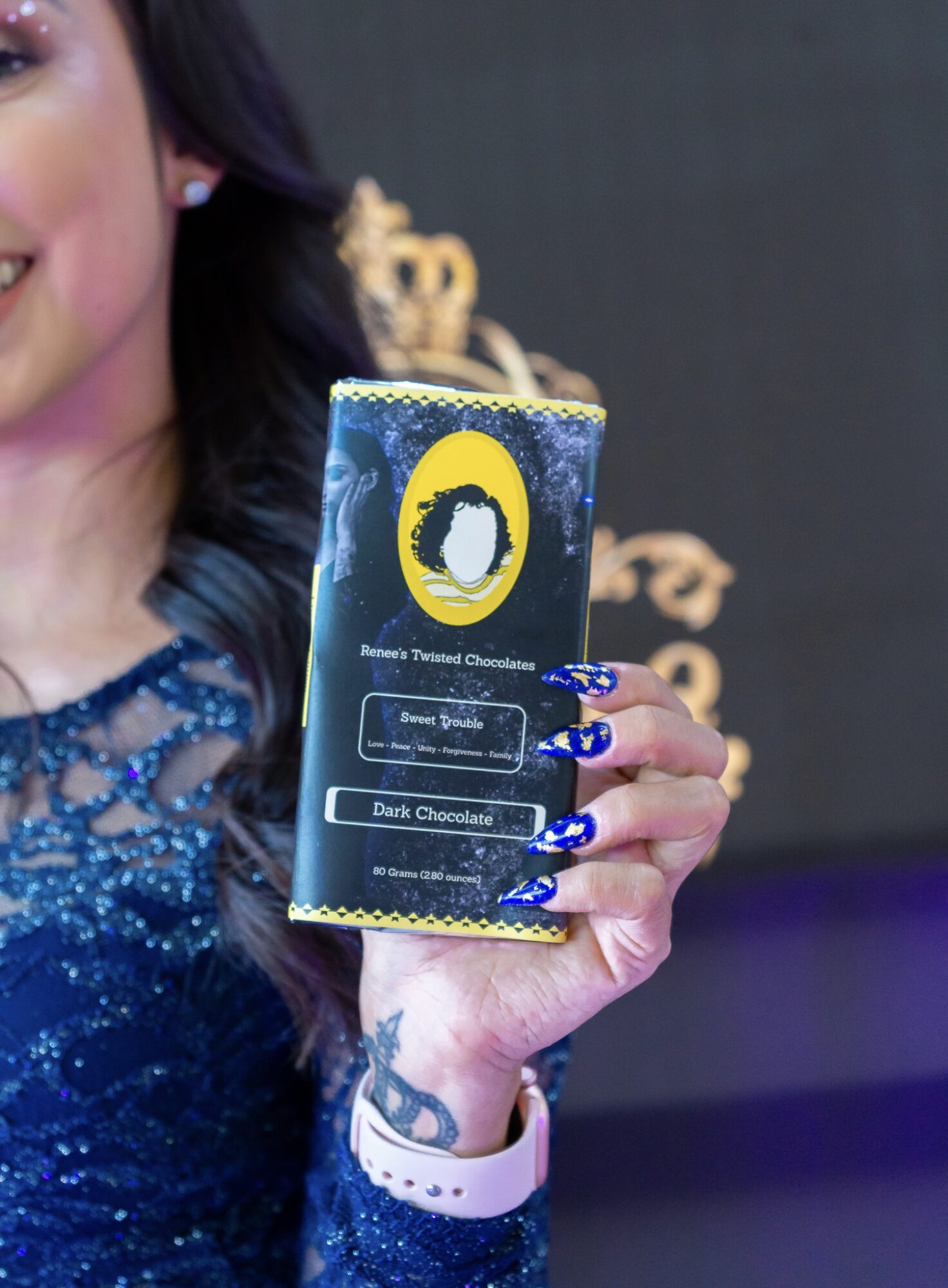 Person holding a dark chocolate bar with a blue and yellow label, wearing a blue dress and a white watch.