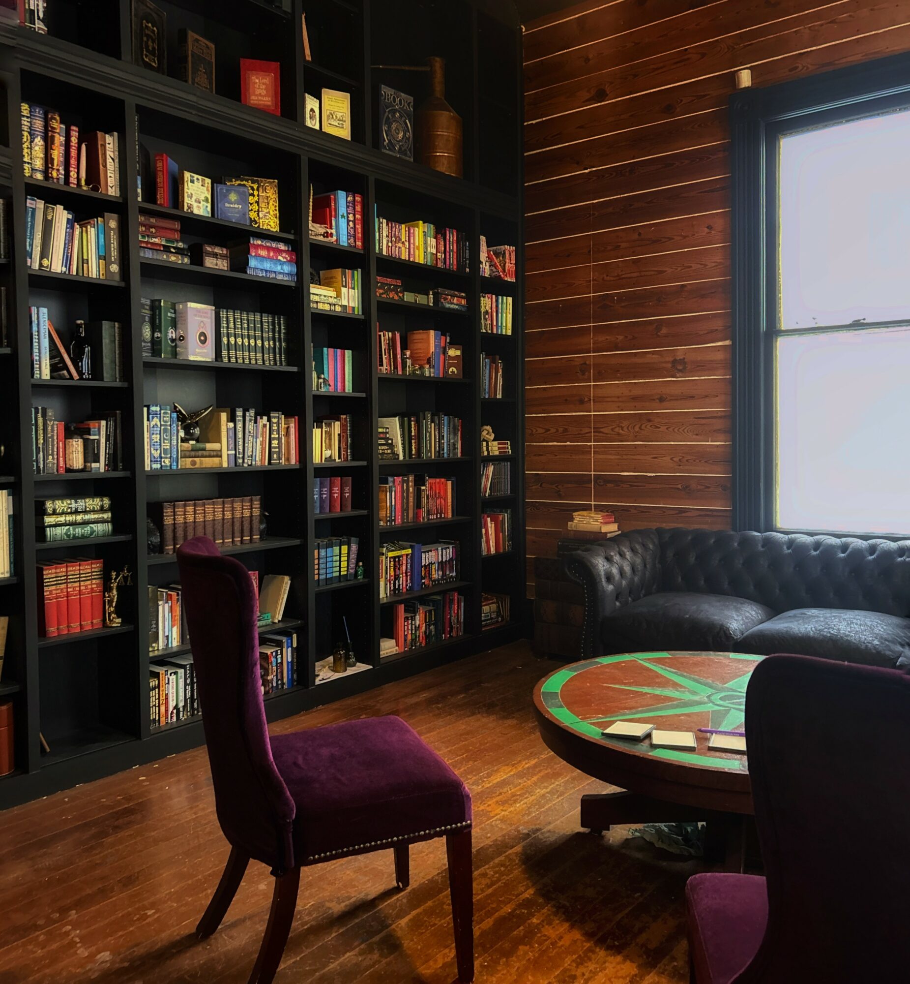 Bookshelf filled with books, a purple chair, a round table, and a window with sunlight, in a cozy room.