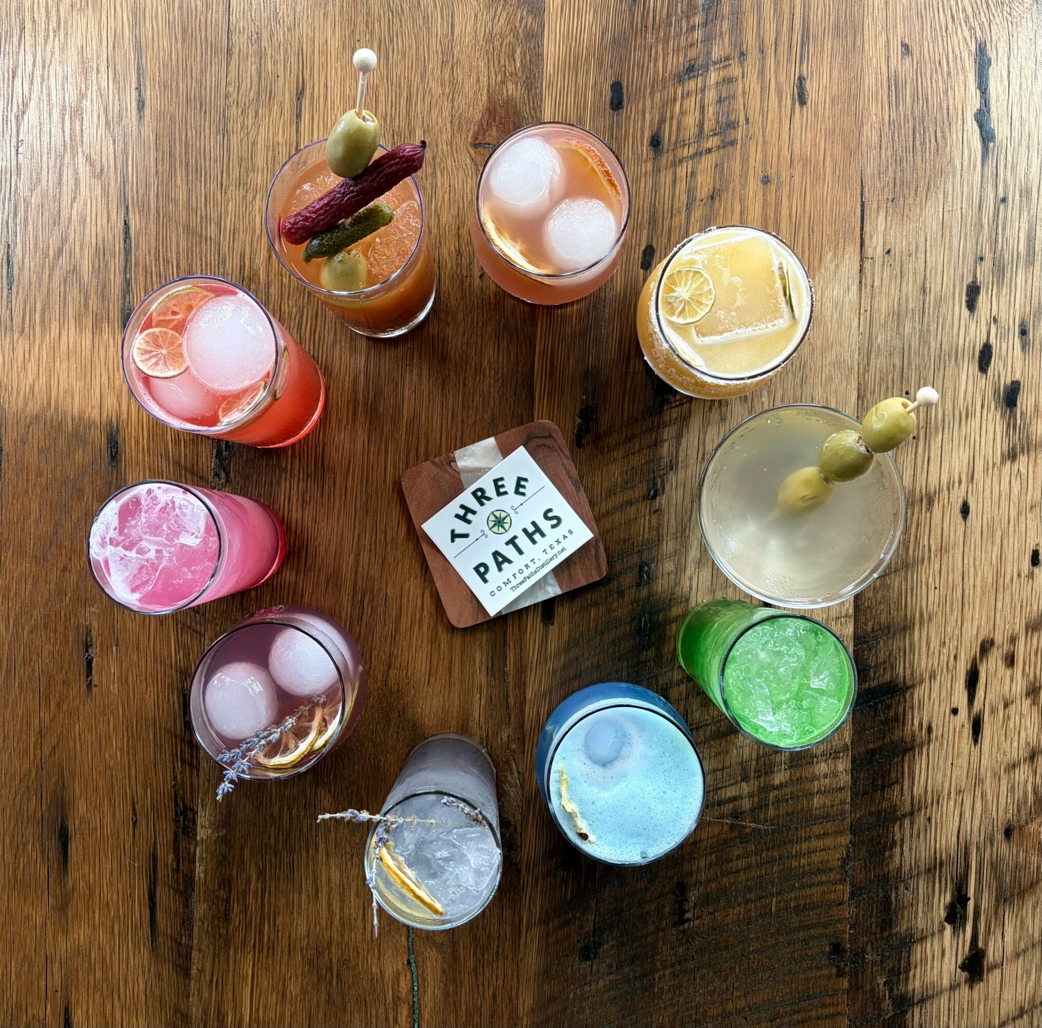 Top-down view of nine colorful cocktails with garnishes on a wooden table, with a small card reading 'Three Paths'.