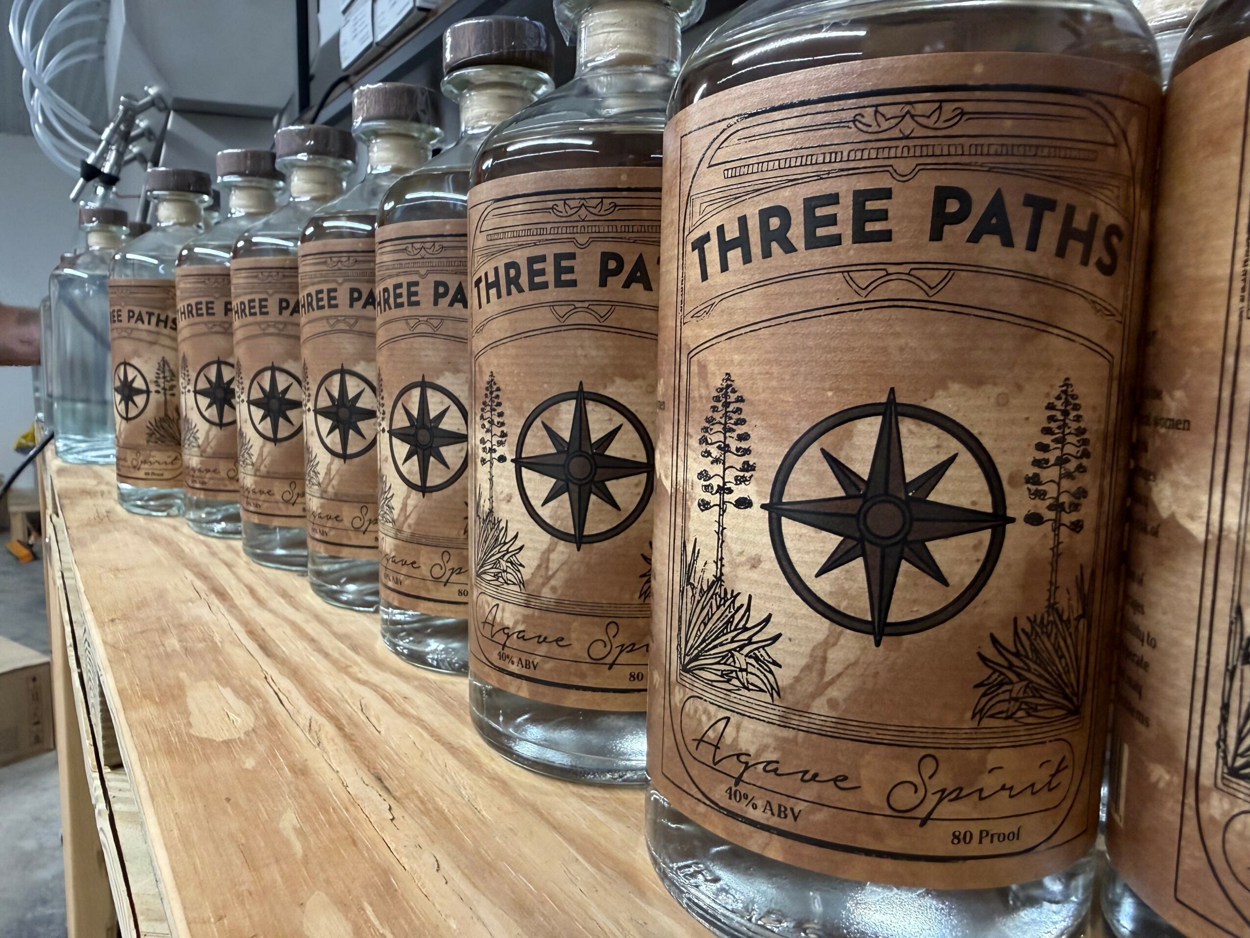 Row of glass bottles with brown paper labels reading 'Three Paths' and a compass design, on a wooden surface.