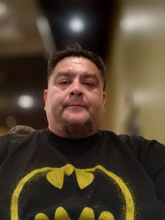 Man with short dark hair and goatee wearing a black Batman logo shirt, indoors with blurred background.