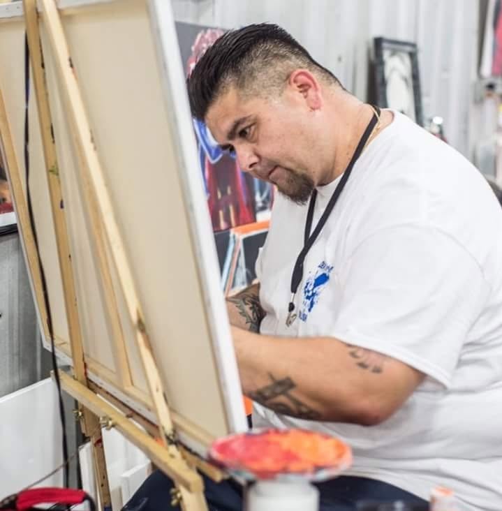 Man painting on canvas, focused, in a studio, with art supplies nearby, wearing a white shirt, tattoos visible on arm.