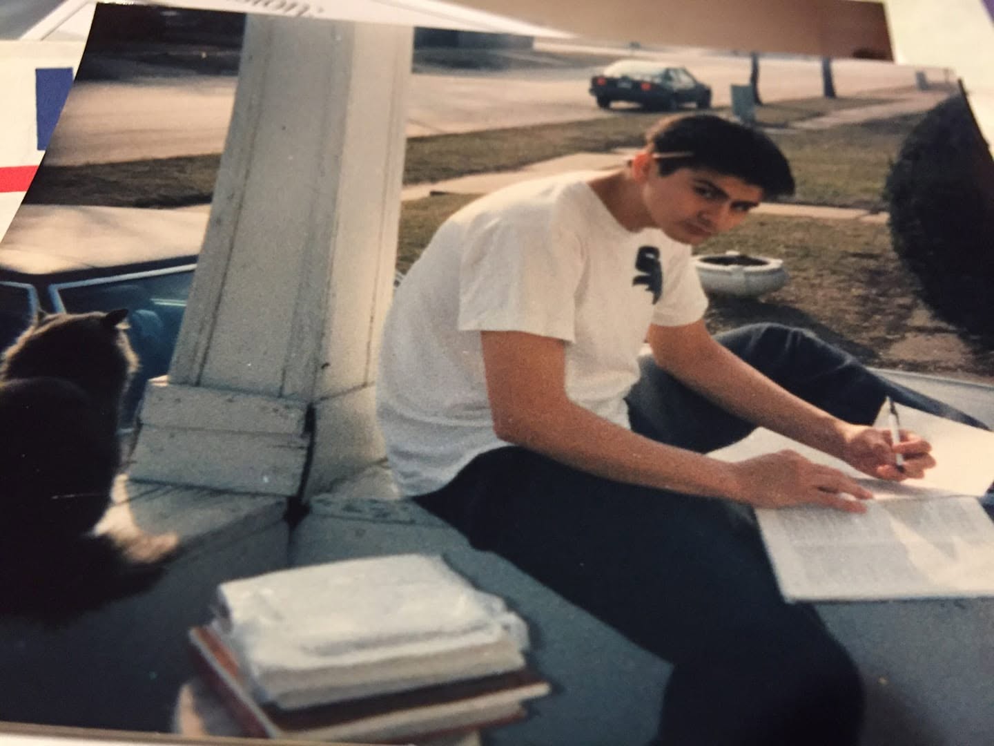 Young person sitting outdoors with papers, a stack of books, and a cat nearby, during daytime.