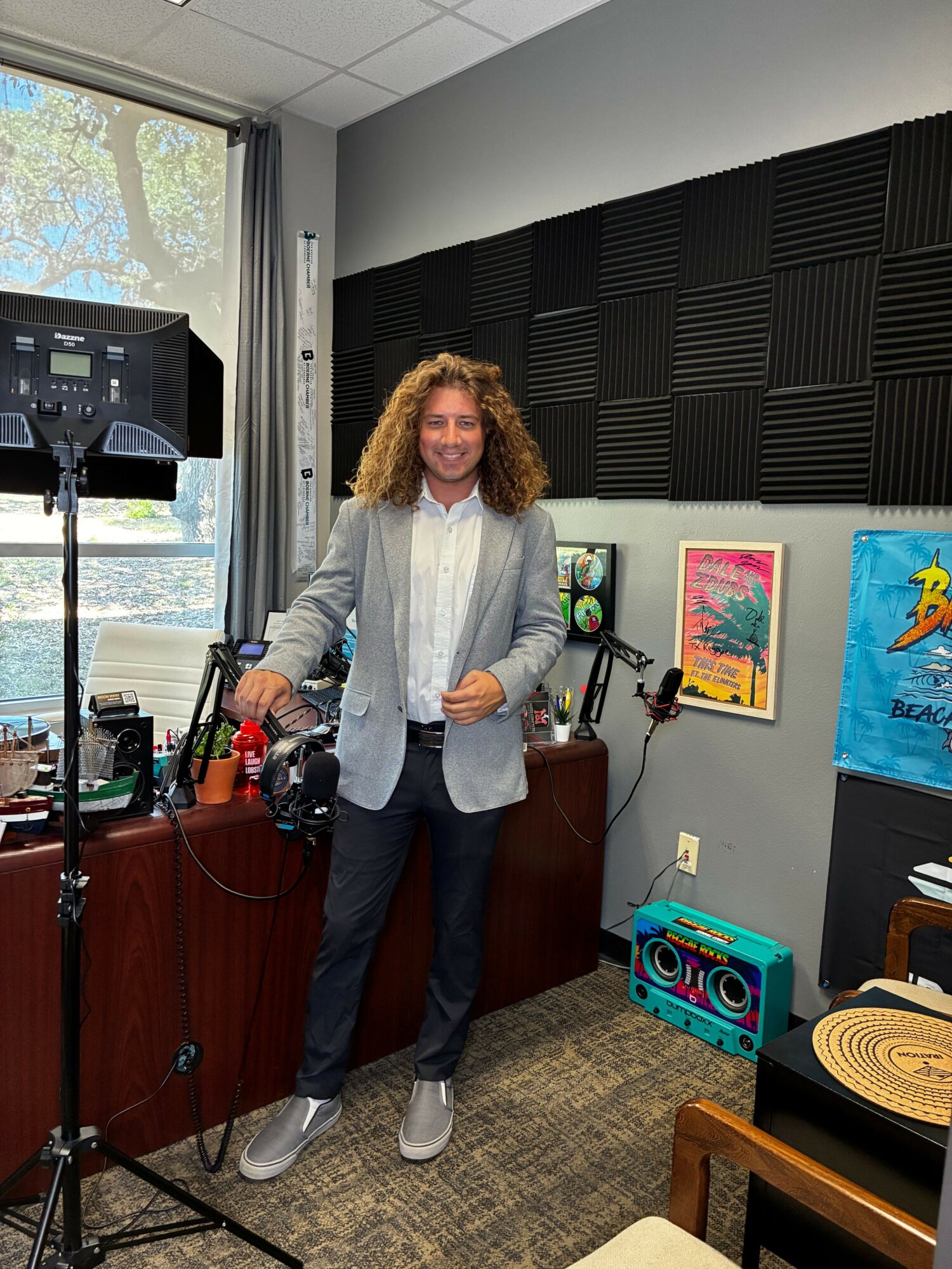 Person with curly hair standing in a room with artwork and audio equipment, holding a microphone, facing camera.