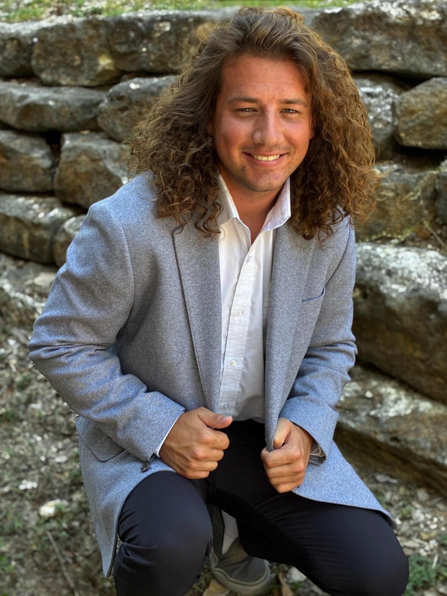 Man with long curly hair smiling, wearing a light gray blazer and white shirt, sitting outdoors in front of rocks.