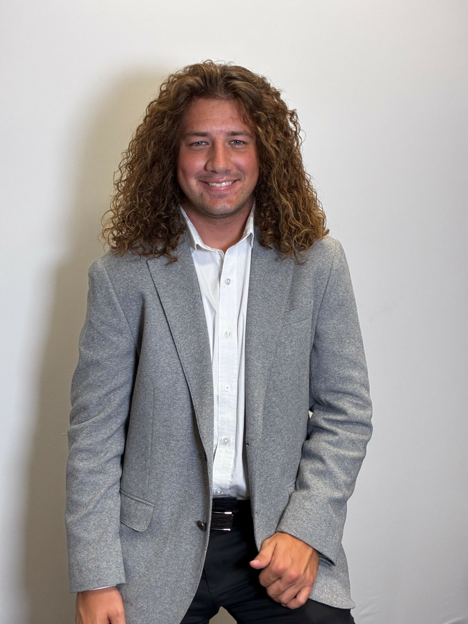 Woman with curly hair wearing a gray blazer and white shirt, smiling, standing against a plain background.