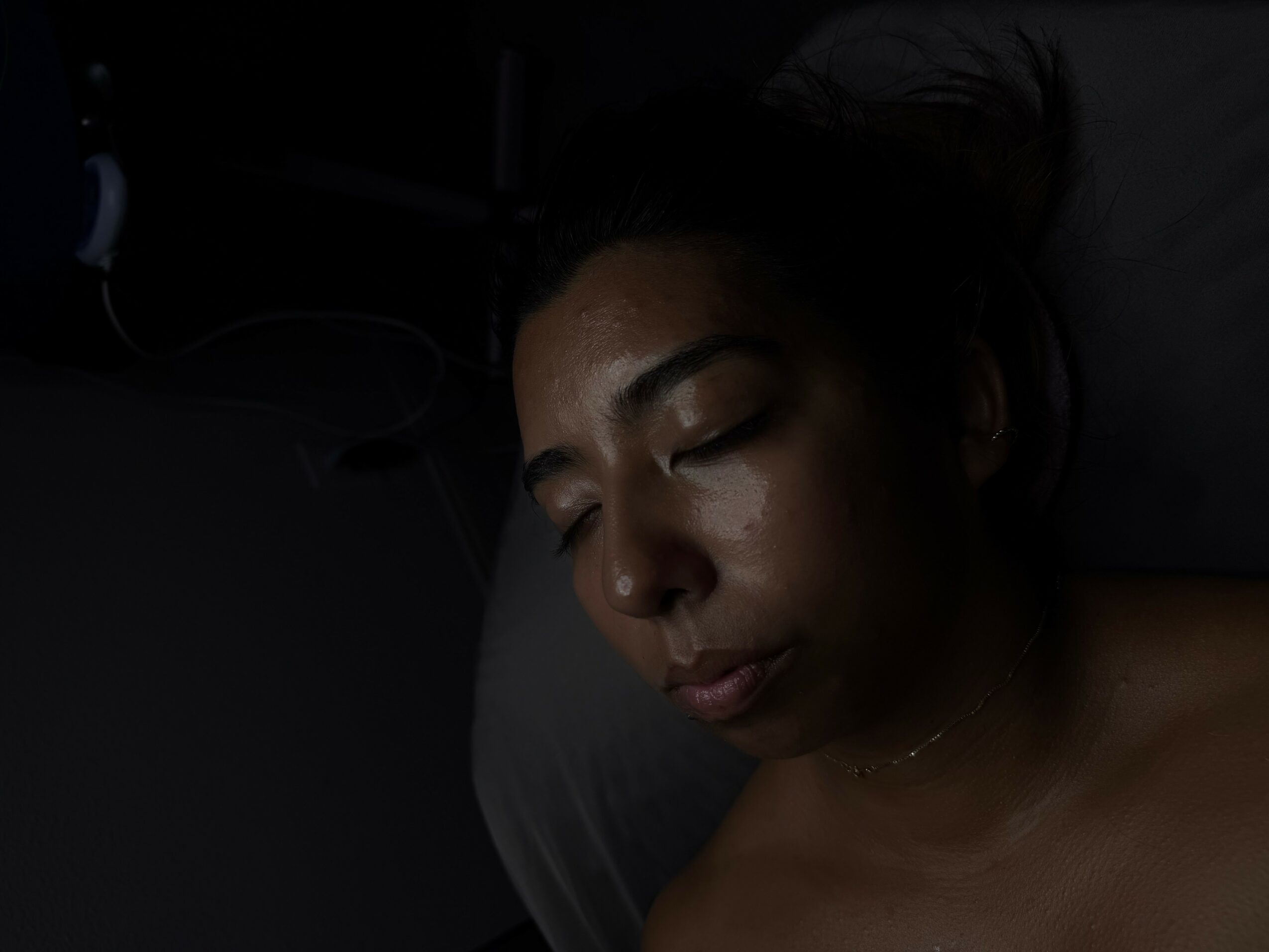 Person sleeping with eyes closed on dark pillow, face illuminated slightly.