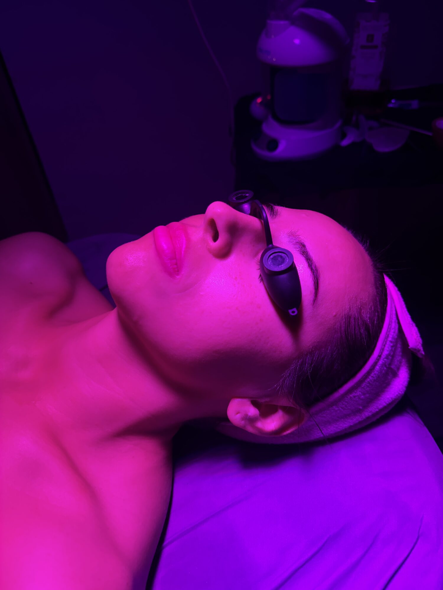 Woman lying in bed wearing a sleep mask, illuminated by purple light, with a serious expression.