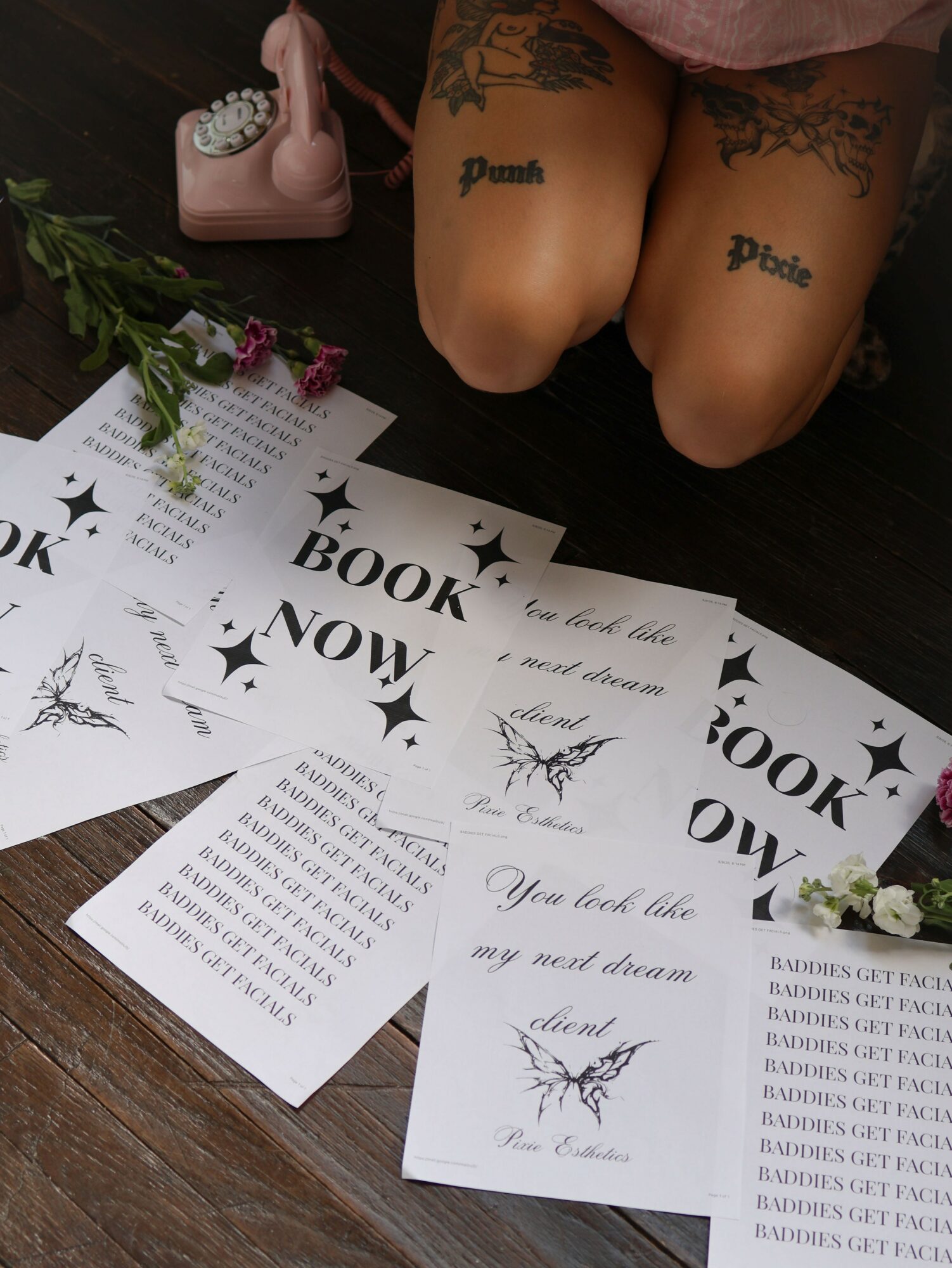 Person kneeling on floor with tattoos on thighs, surrounded by papers and a pink telephone, with flowers nearby.