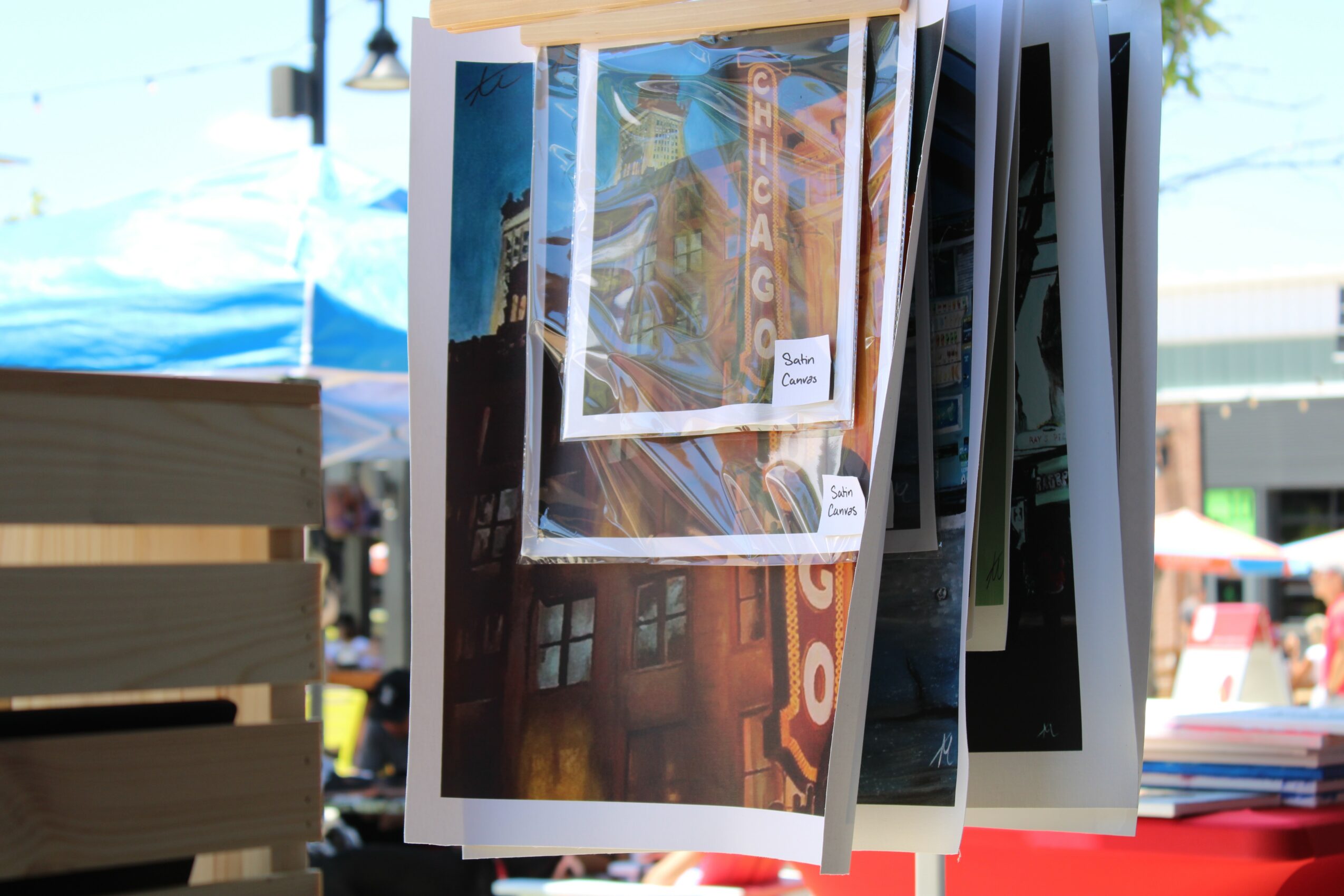 Photographs hanging on display at an outdoor market, with tents and people in the background.