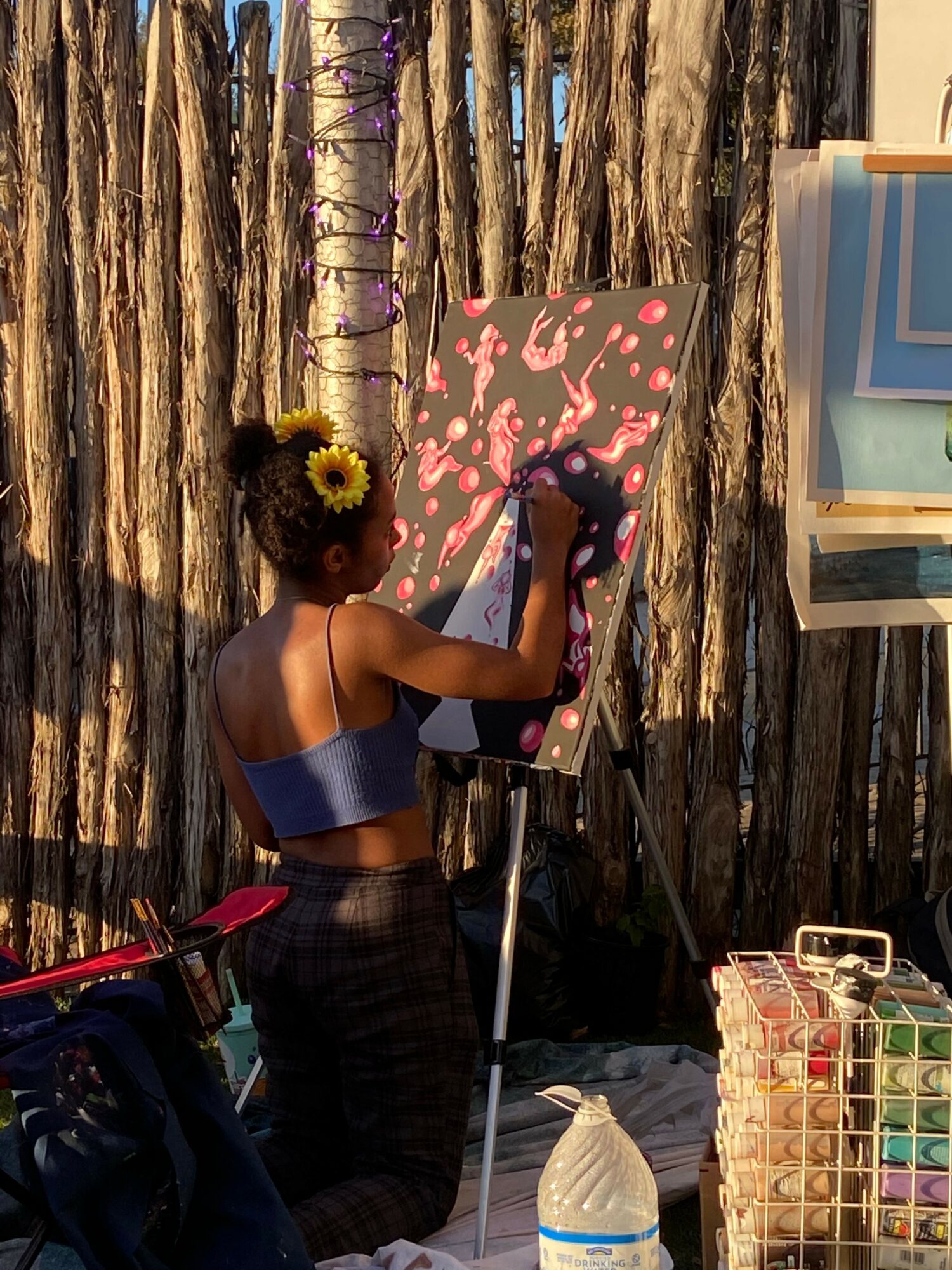 Person with curly hair and sunflower headband holding a pink and black patterned umbrella, standing outdoors near wooden logs.