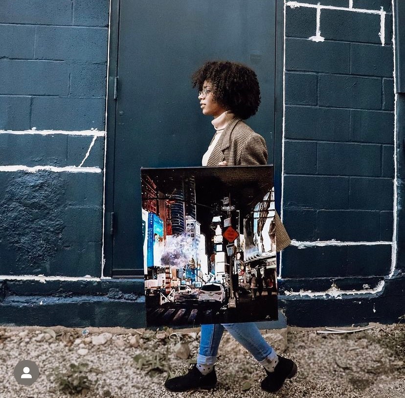 Person walking along a wall carrying a cityscape painting, wearing a coat and jeans, with a textured ground and dark wall background.