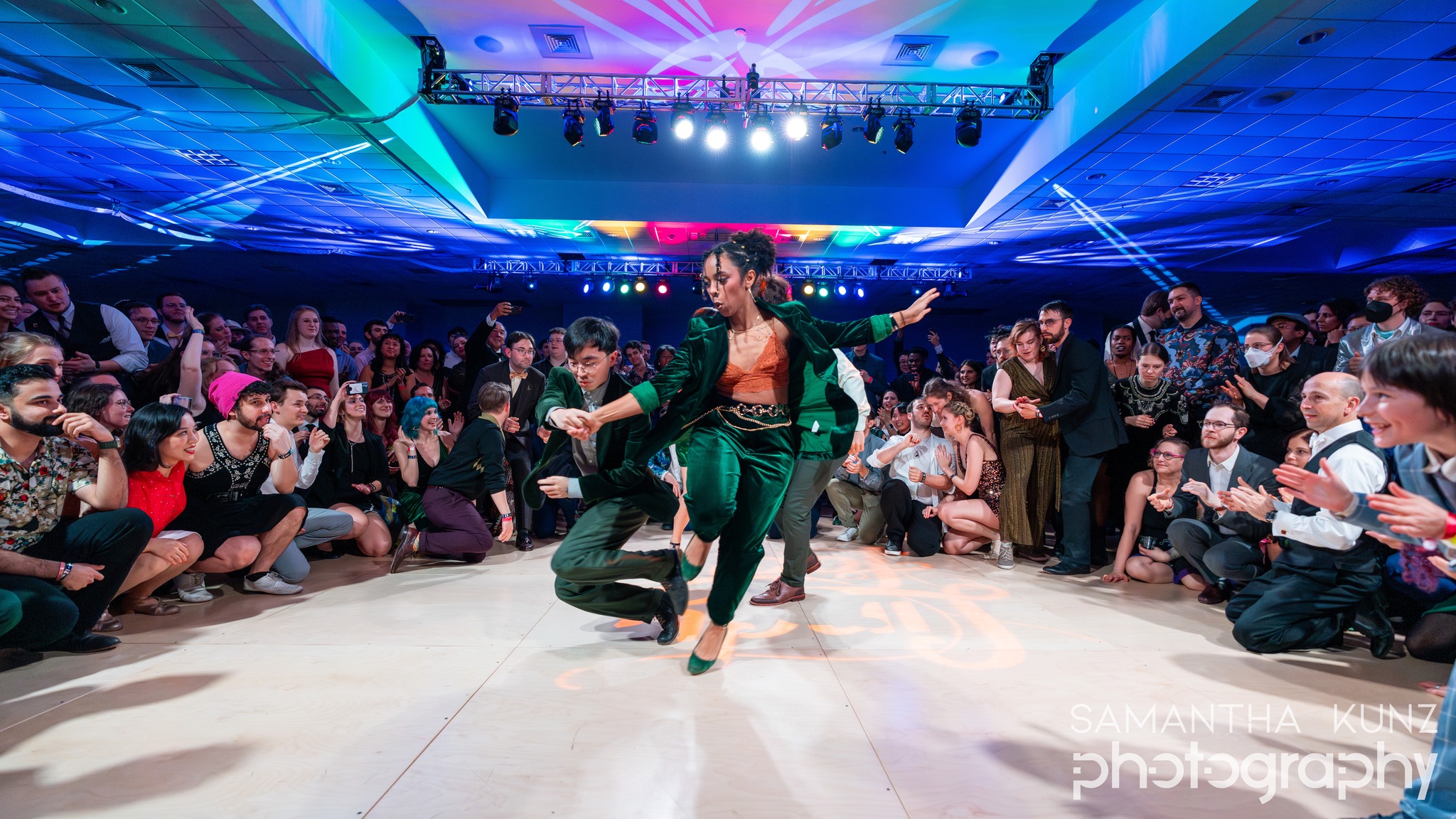 Dancer in green costume performing in the center of a large audience at an indoor event with colorful lighting