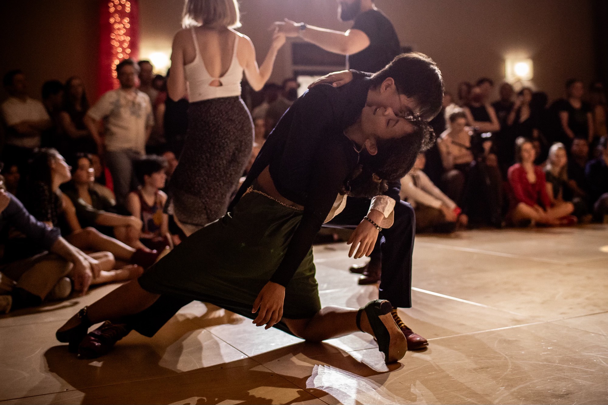 Person in black clothing crouches on the dance floor with a crowd watching in the background.