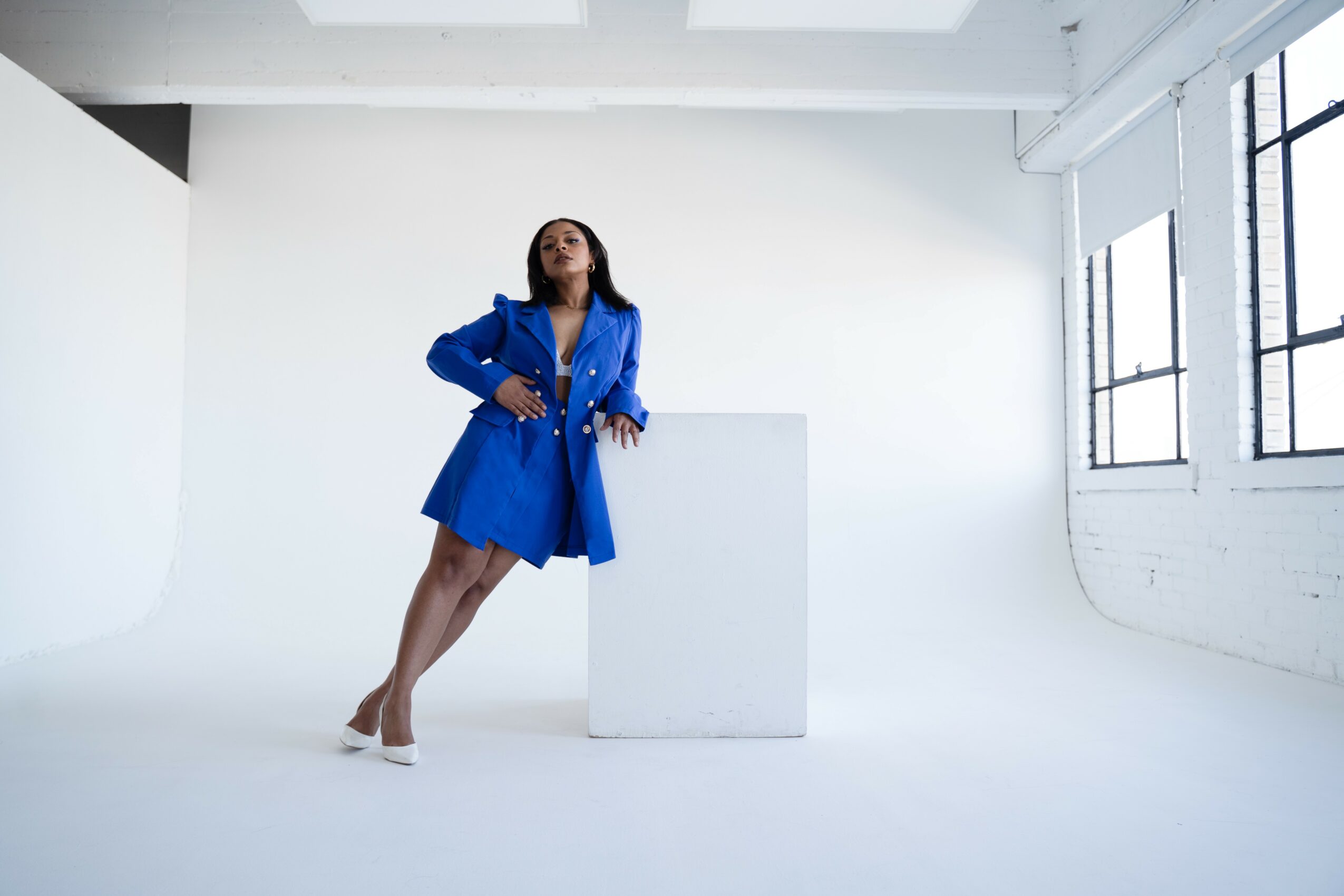 Woman in blue coat leaning on a white pedestal in a bright, minimal studio with large windows.