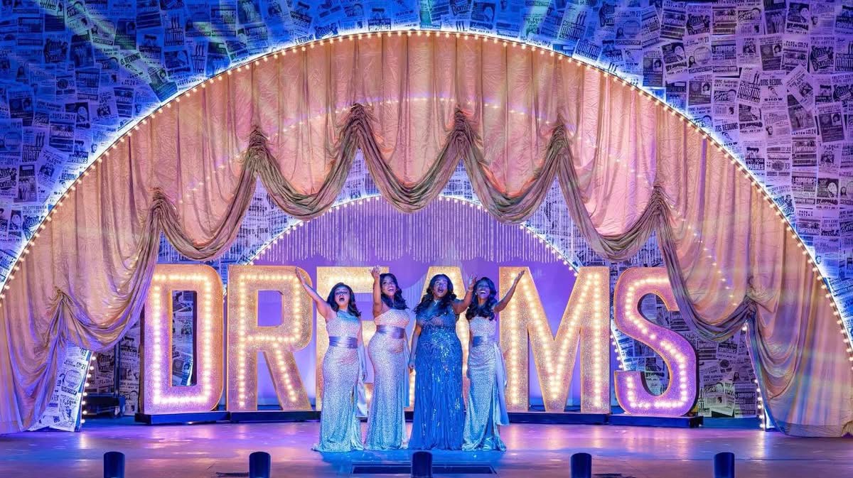 Four women in elegant dresses stand in front of large illuminated letters spelling 'DREAMS' on a decorated stage.