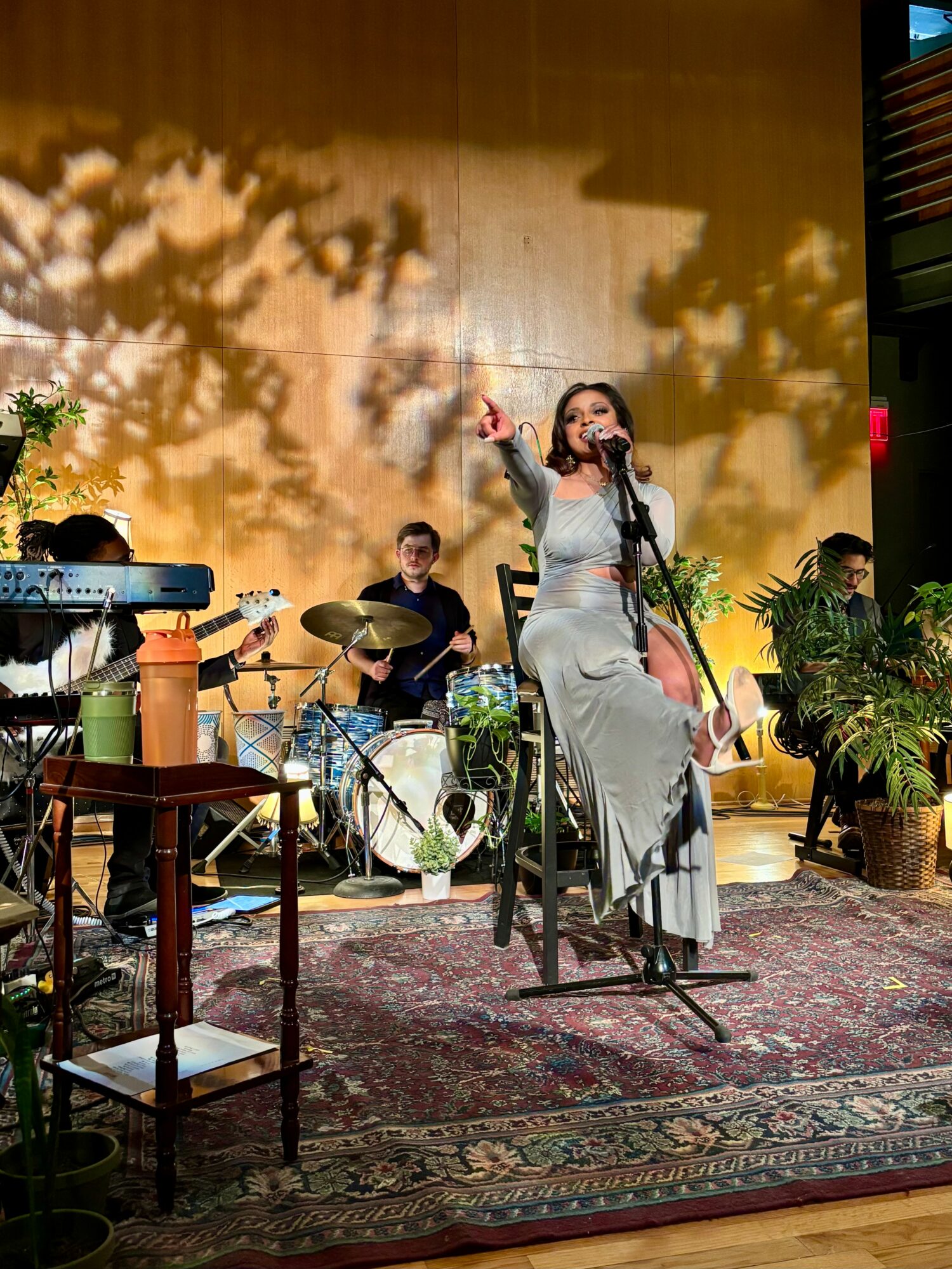 A woman singing into a microphone on stage with musicians, plants, and a patterned rug, illuminated by warm lighting.