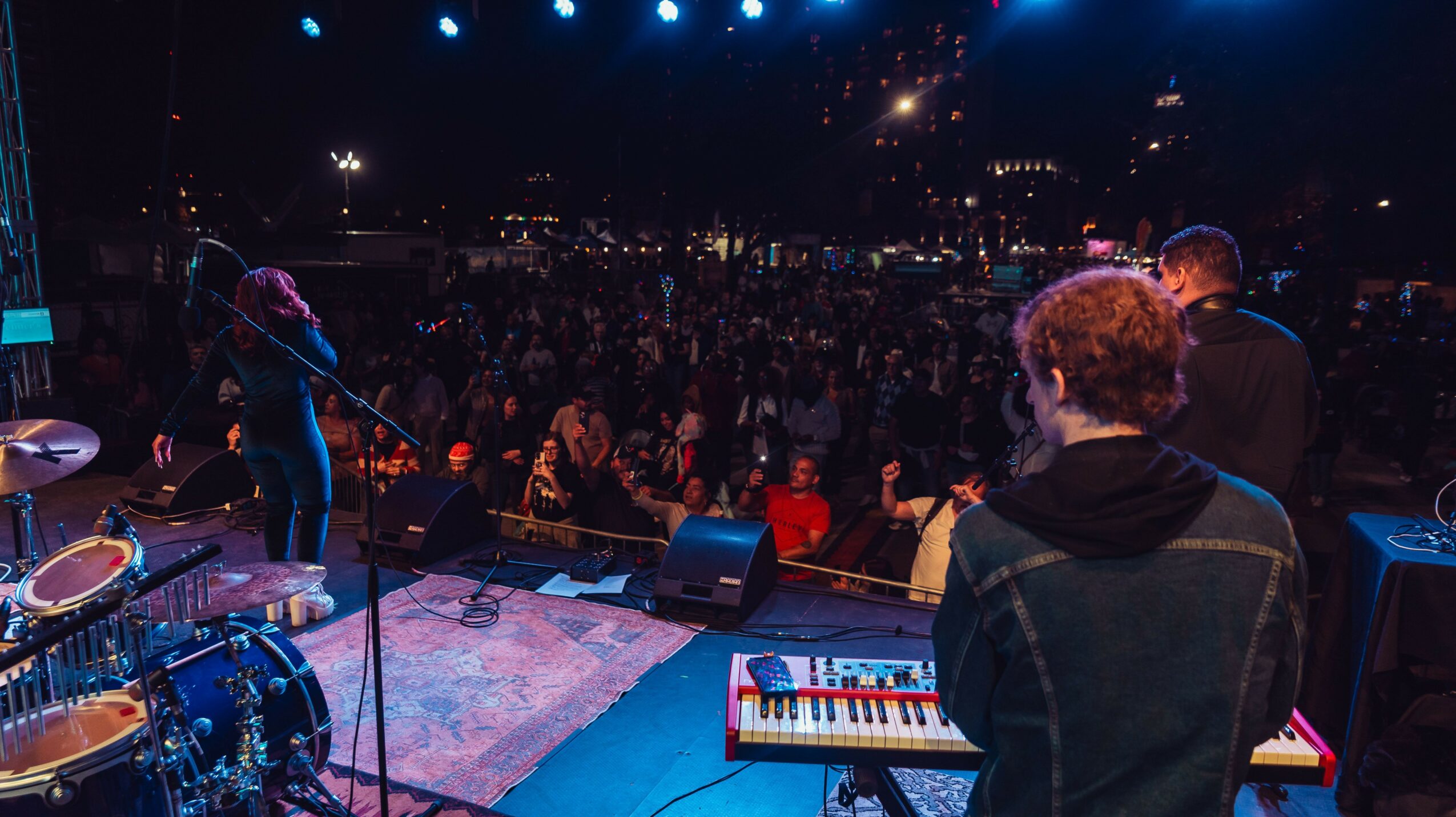 Musicians perform on stage with a crowd in the background at night, stage equipment visible, audience watching.