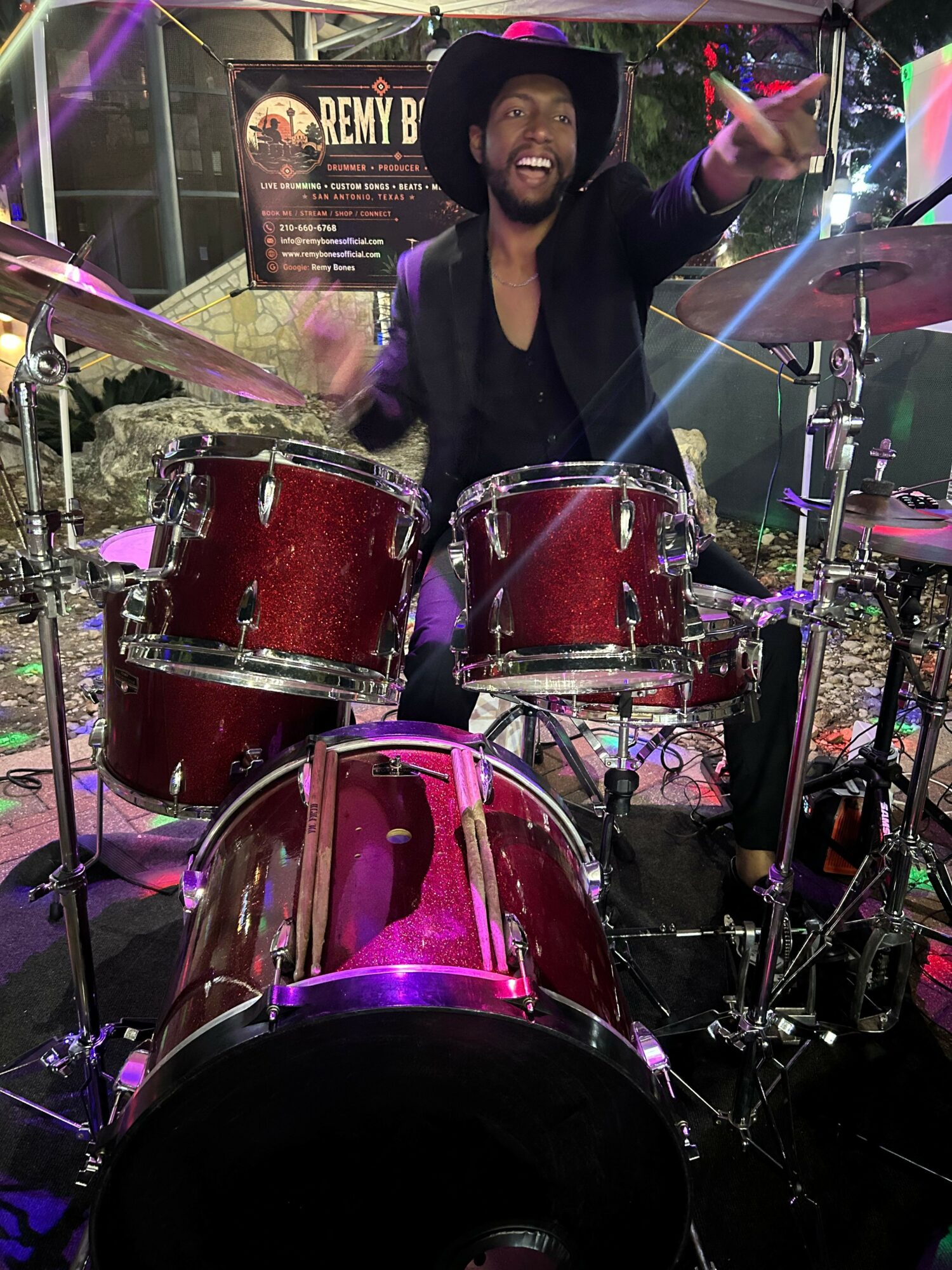 Person playing drums on stage with colorful lighting and musical equipment, smiling and raising a hand.