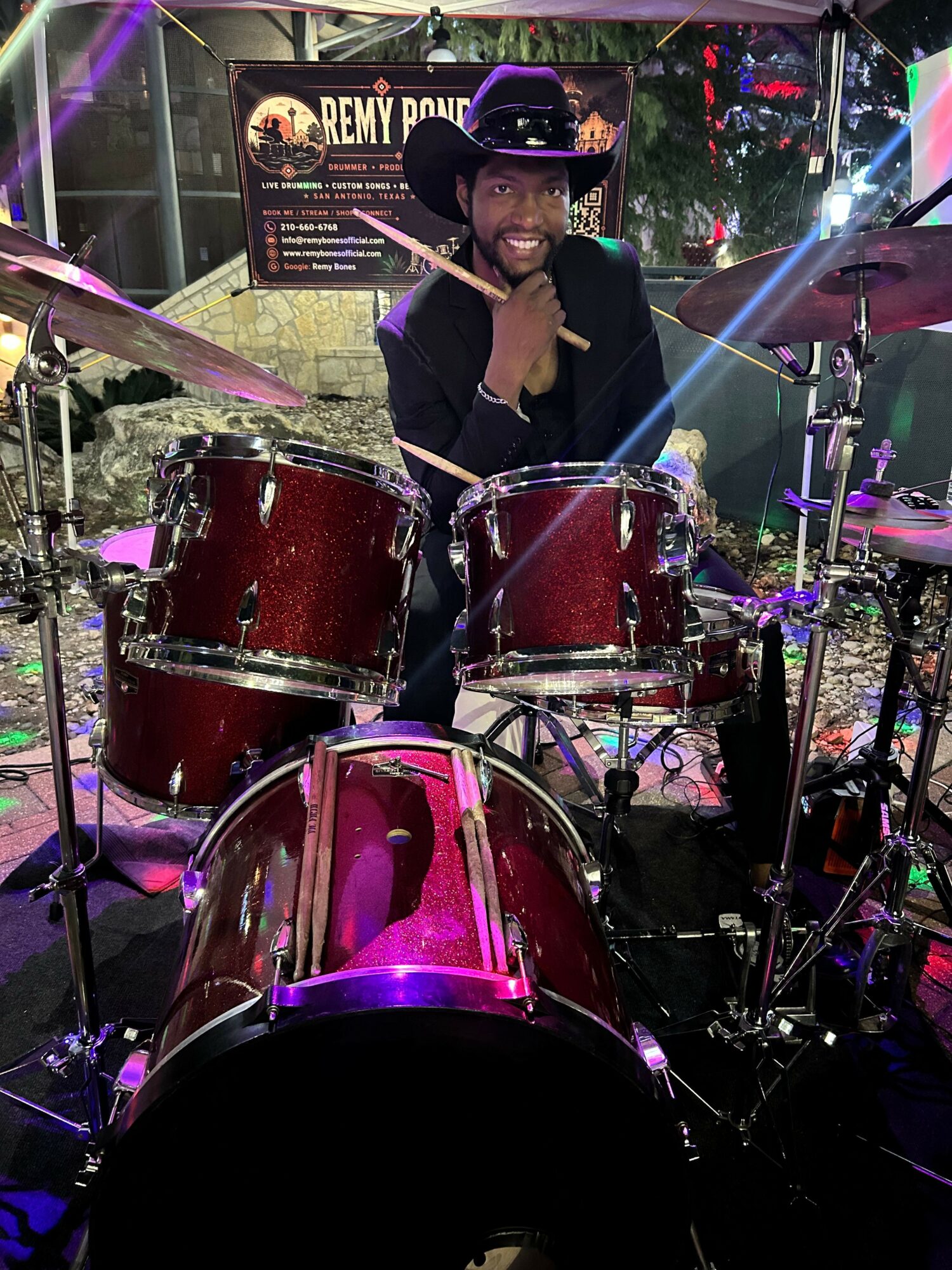 Person playing drums on stage with colorful lights and equipment, audience visible in background.