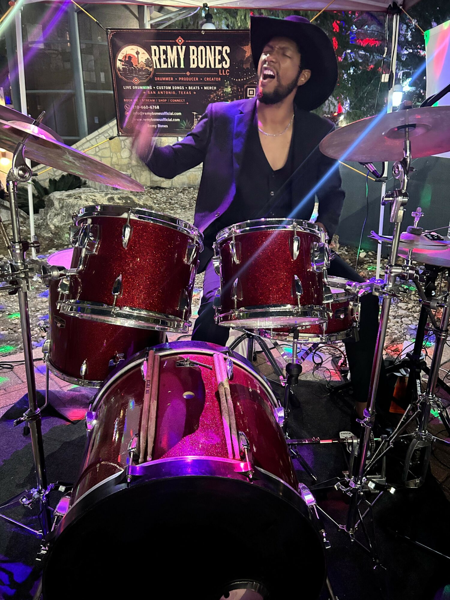 A woman with long dark hair playing drums on stage with colorful lights and a sign in the background.