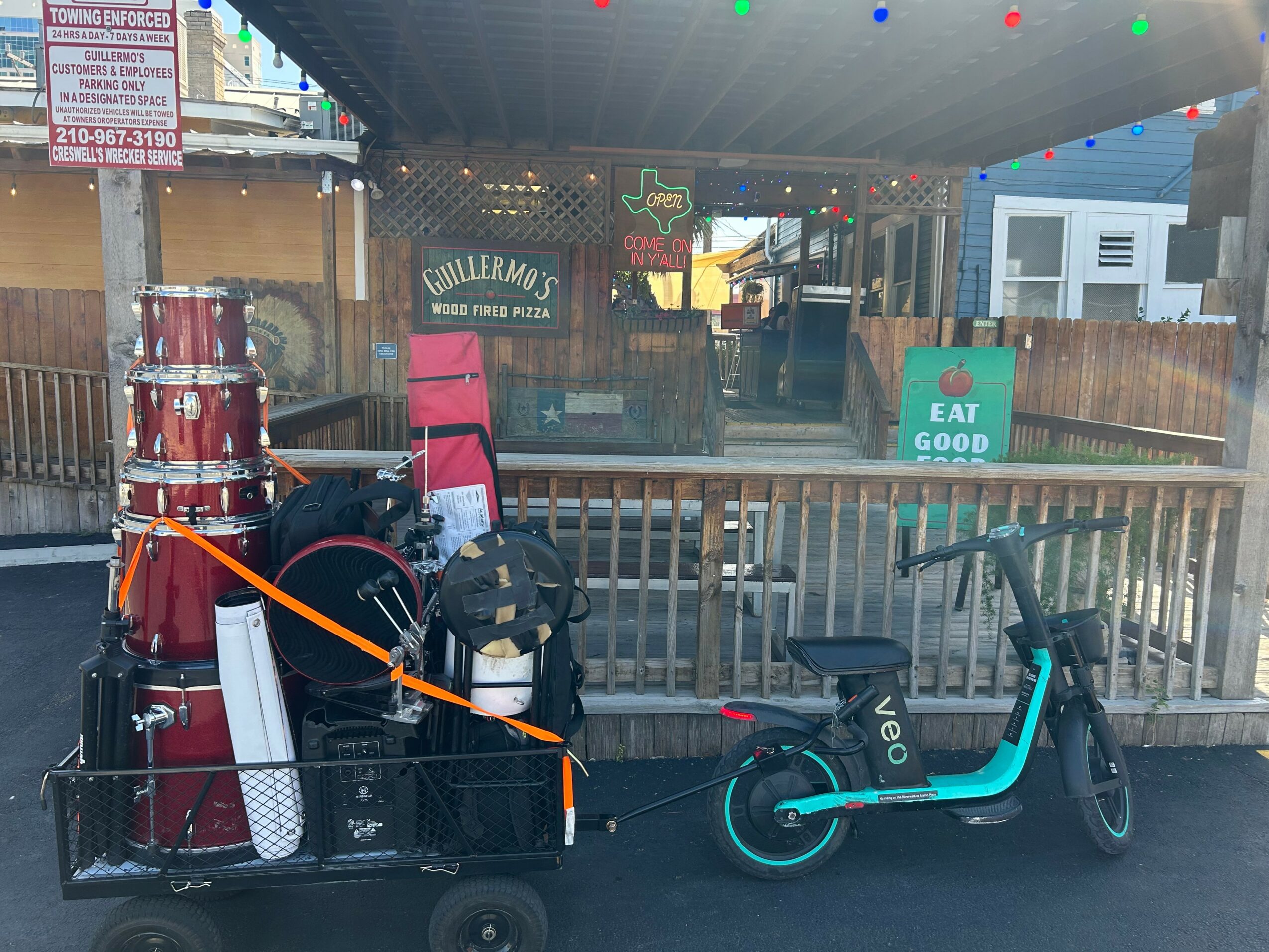 A cart with drums and gear parked next to a blue electric scooter outside a fenced area with a sign that says 'EAT GOOD'.