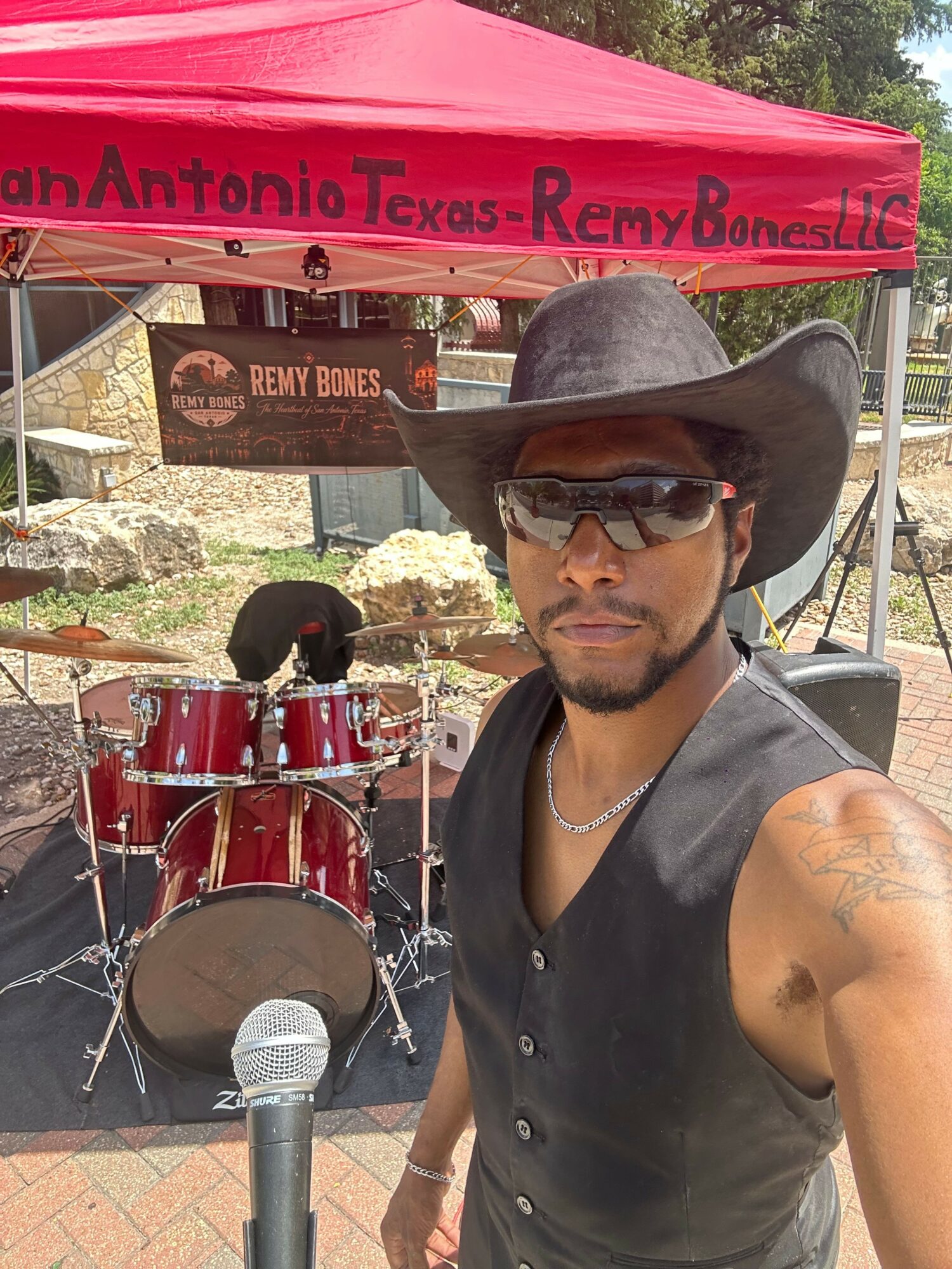 Man wearing a cowboy hat, sunglasses, and sleeveless shirt, standing outdoors with a drum set and microphone nearby.
