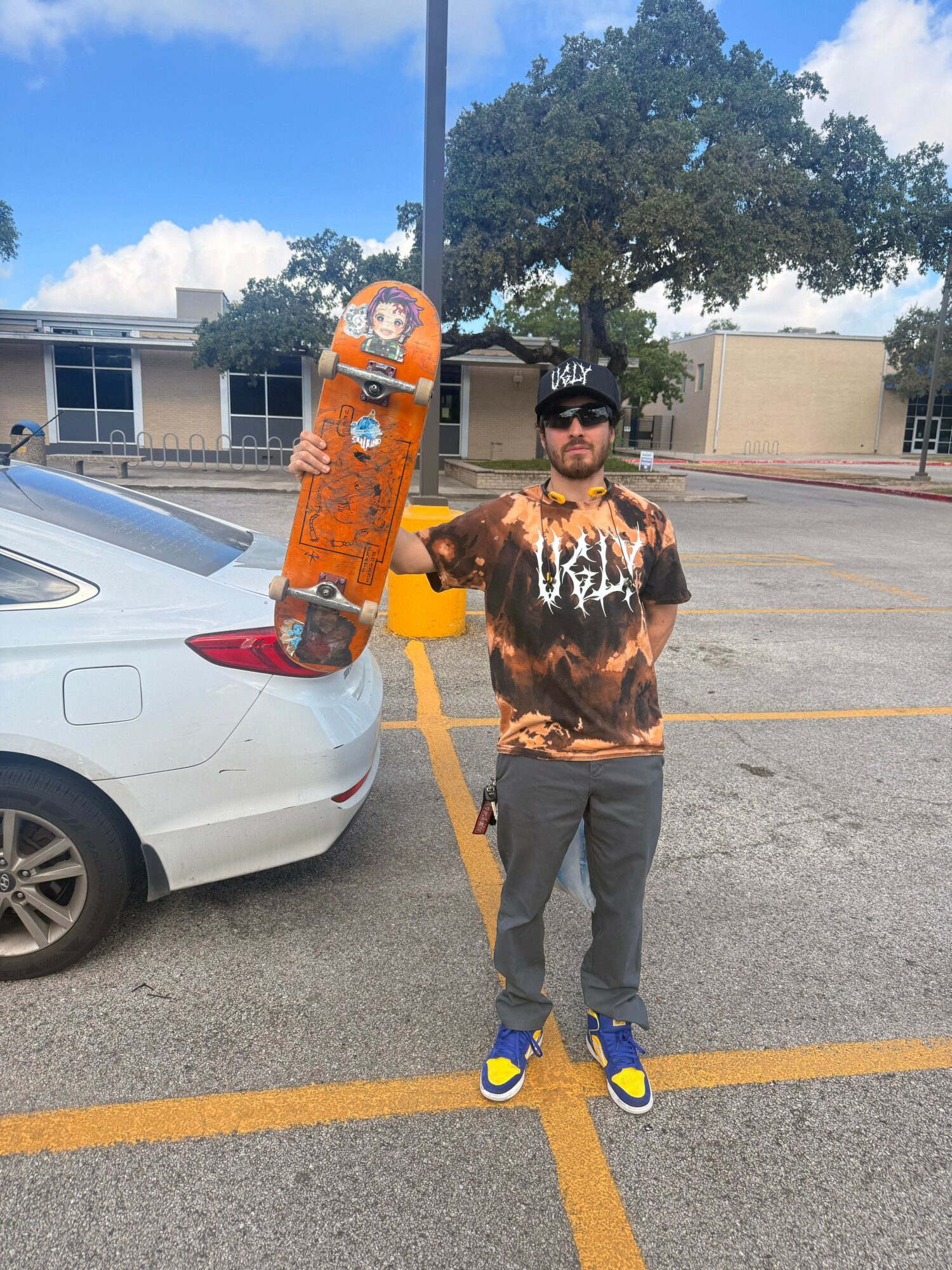 Person standing in parking lot with trees and building in background, wearing a tie-dye hoodie, gray pants, and blue shoes.
