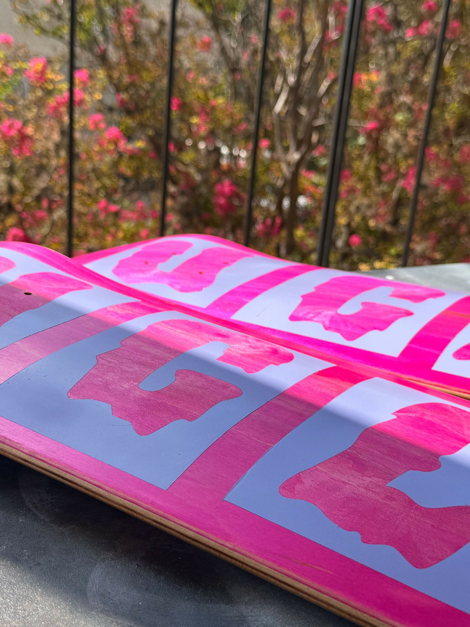 Close-up of a pink and purple sign with text, outdoors with trees and pink flowers in the background.