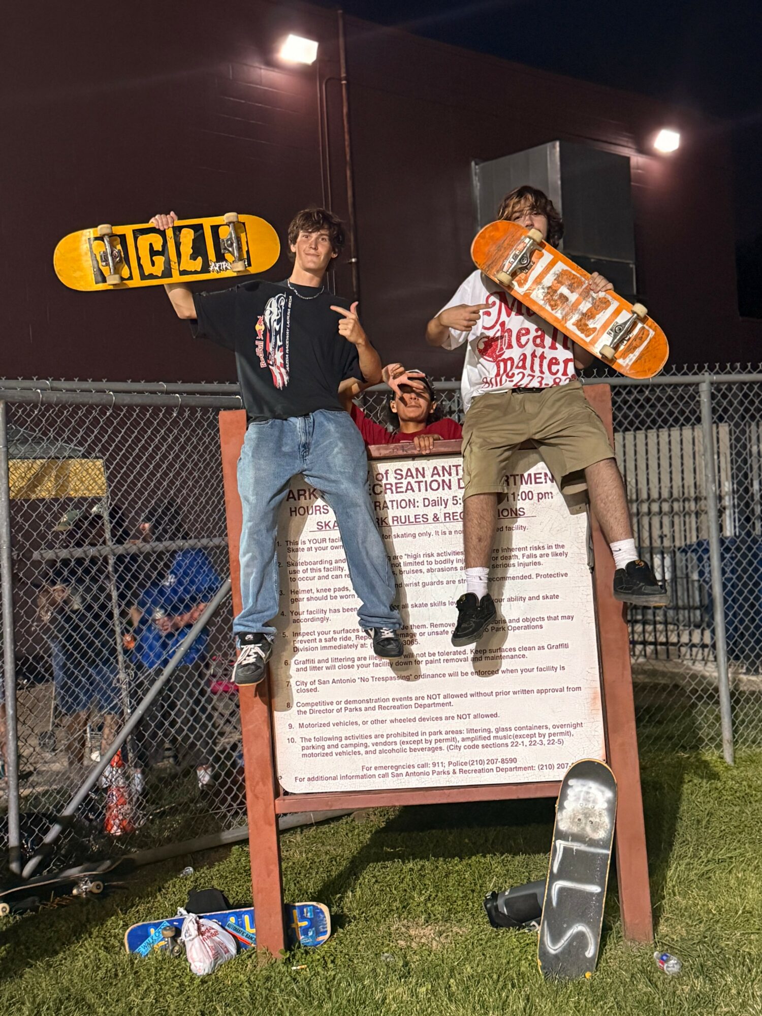 Two young men stand on a signboard holding skateboards, with a fence and night sky in the background.