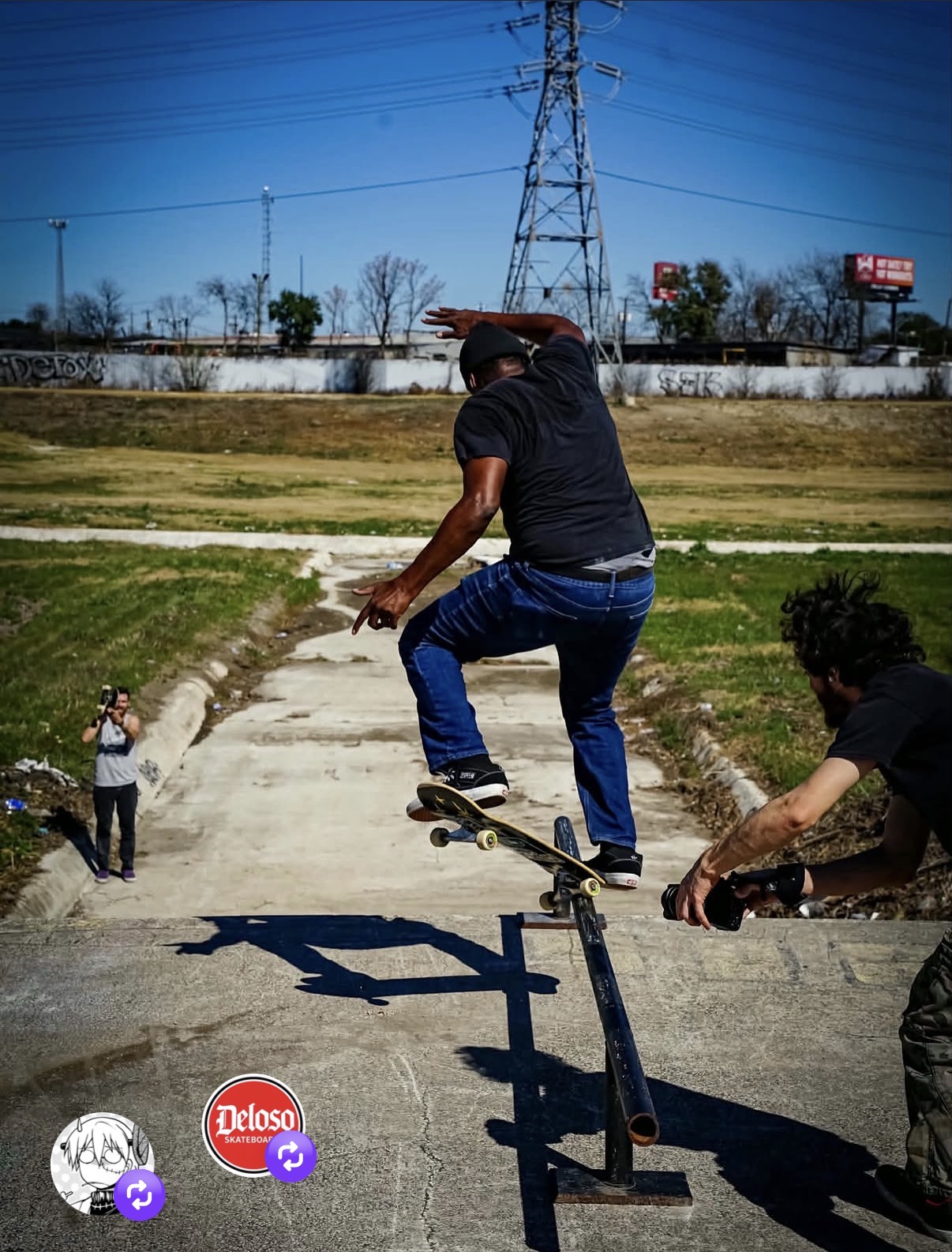 Skateboarder in black shirt and jeans performs a trick on a rail, with two people nearby, outdoors on a sunny day.