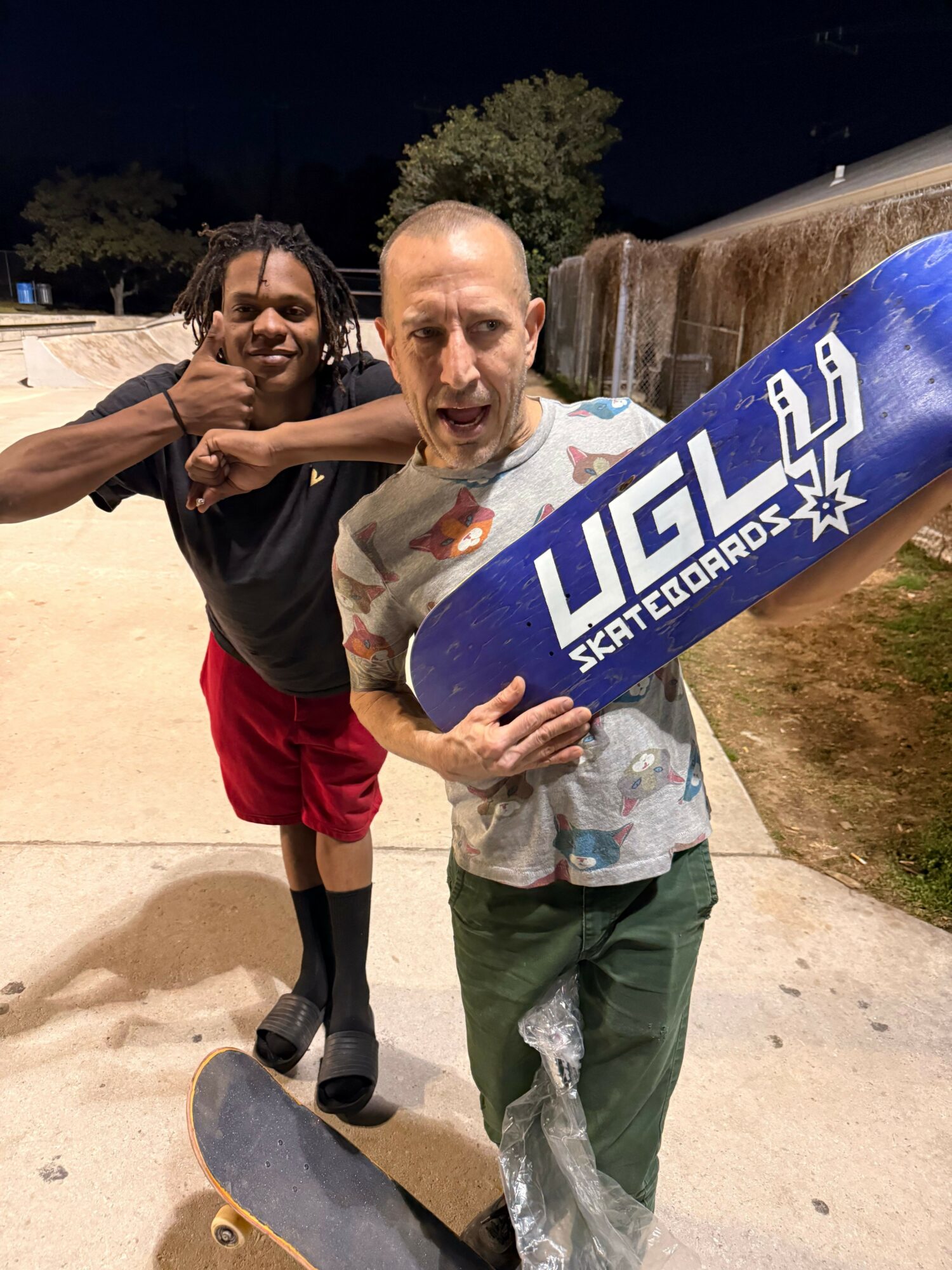 Person holding a skateboard with a blue and white design, standing outdoors at night, with another person nearby.