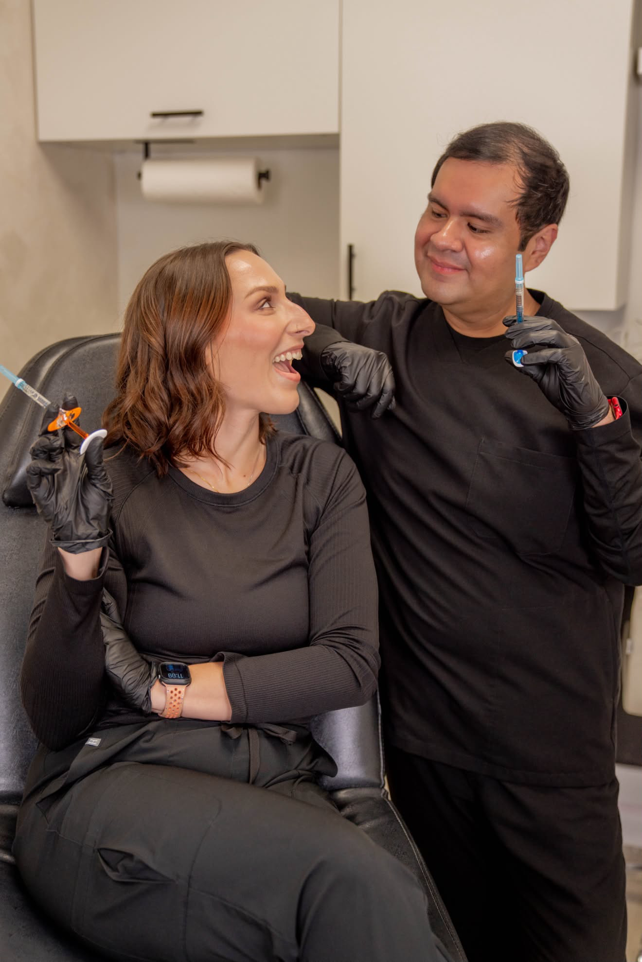 A woman and a man in a medical setting, smiling and talking, with the woman sitting and the man standing, both wearing black clothing and gloves.