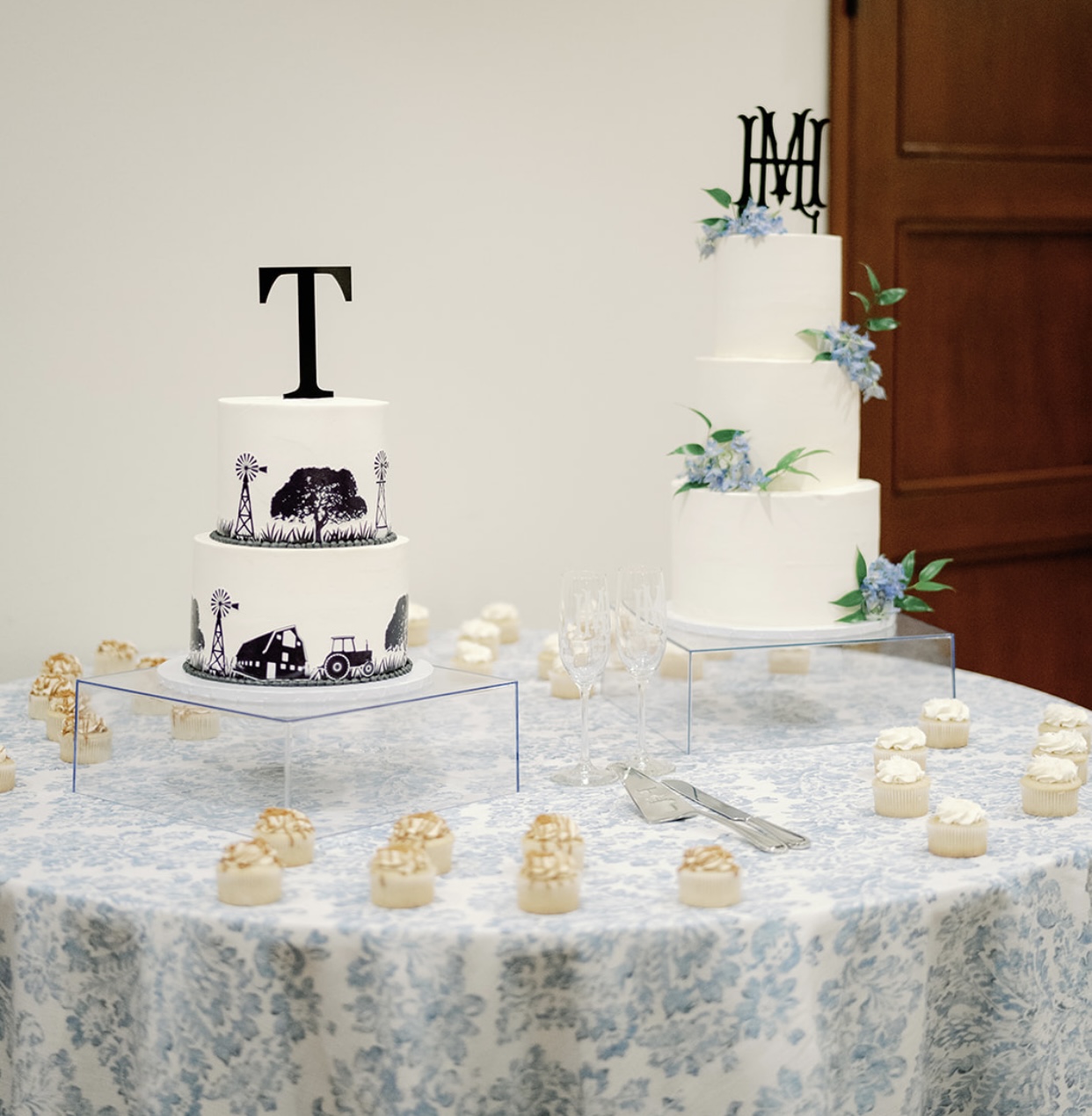Two white tiered cakes with decorations on a table with small desserts, a knife, and a cake server, against a plain wall.