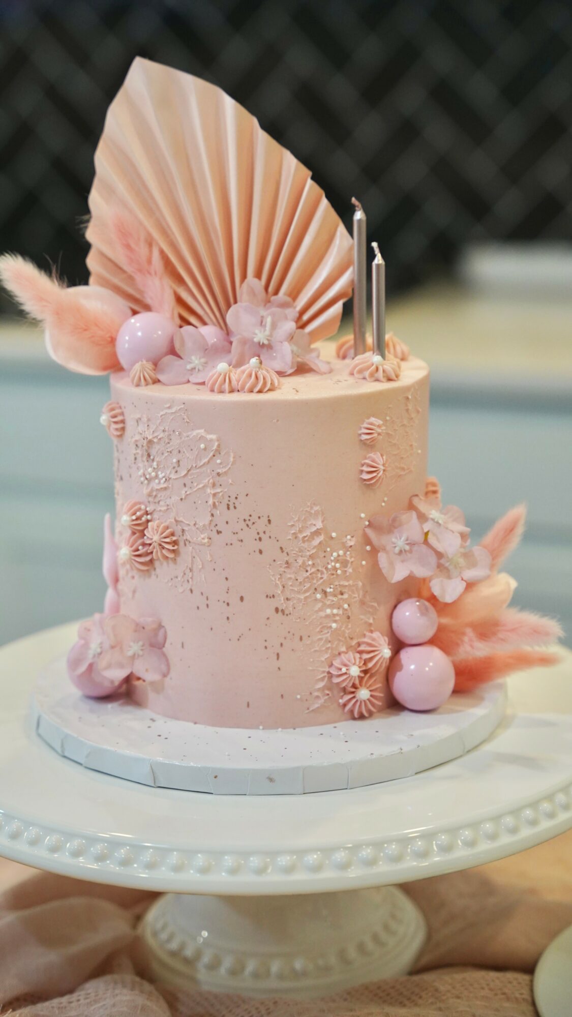 Pink decorated cake with a large fan, flowers, and spherical accents on a white cake stand.