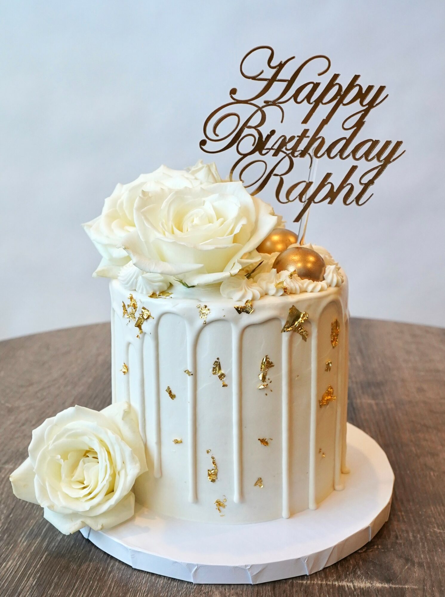 White birthday cake with gold accents, decorated with white roses and a topper reading 'Happy Birthday Ralph'.