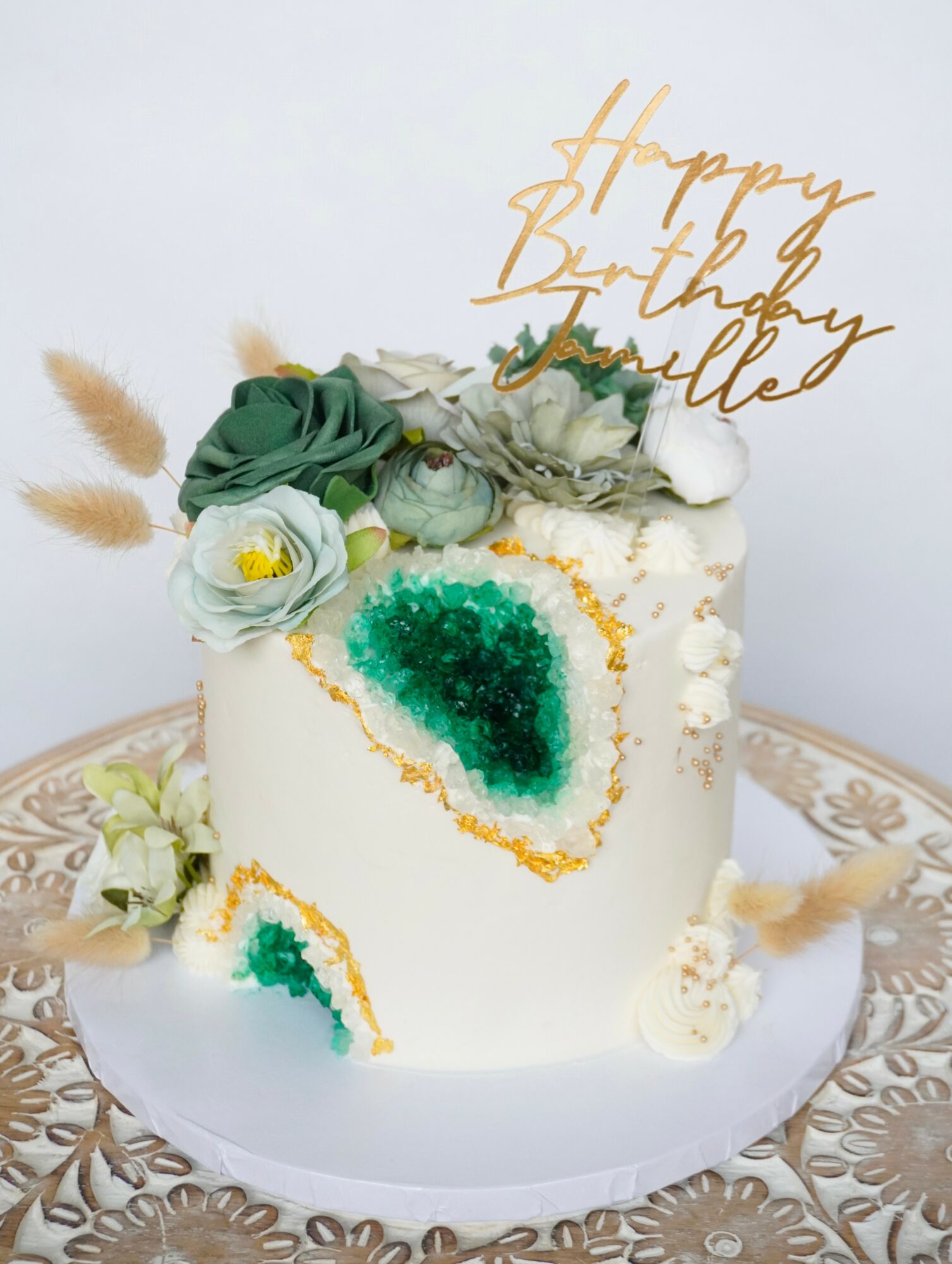 White birthday cake decorated with flowers, green geode, and a gold topper reading 'Happy Birthday'.
