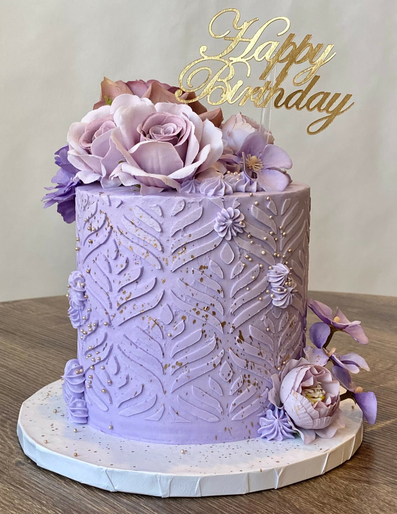 Lavender birthday cake decorated with flowers and a gold topper reading 'Happy Birthday'.