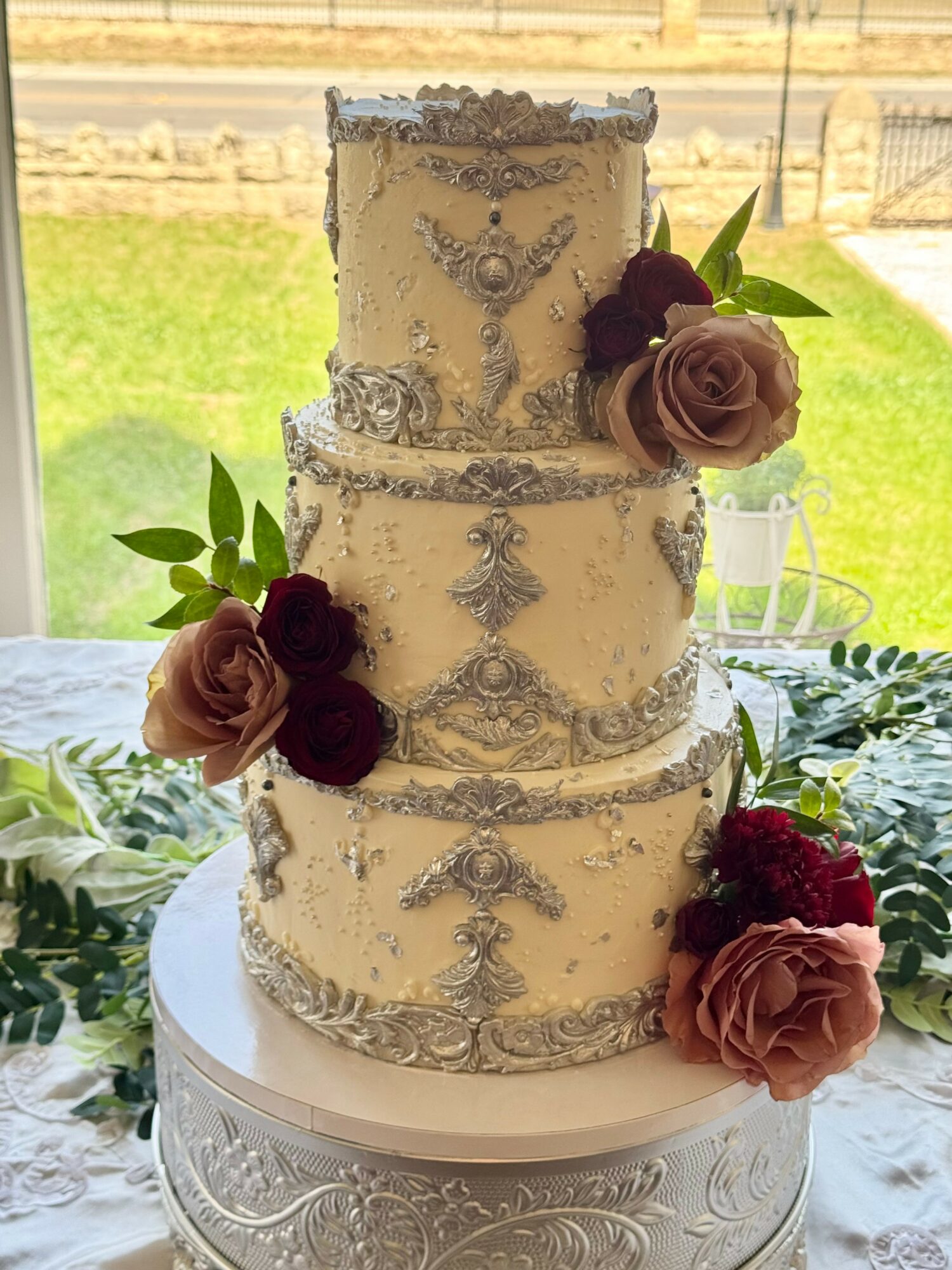 Four-tiered wedding cake decorated with silver embellishments and dark pink and beige roses, placed on a table near a window.