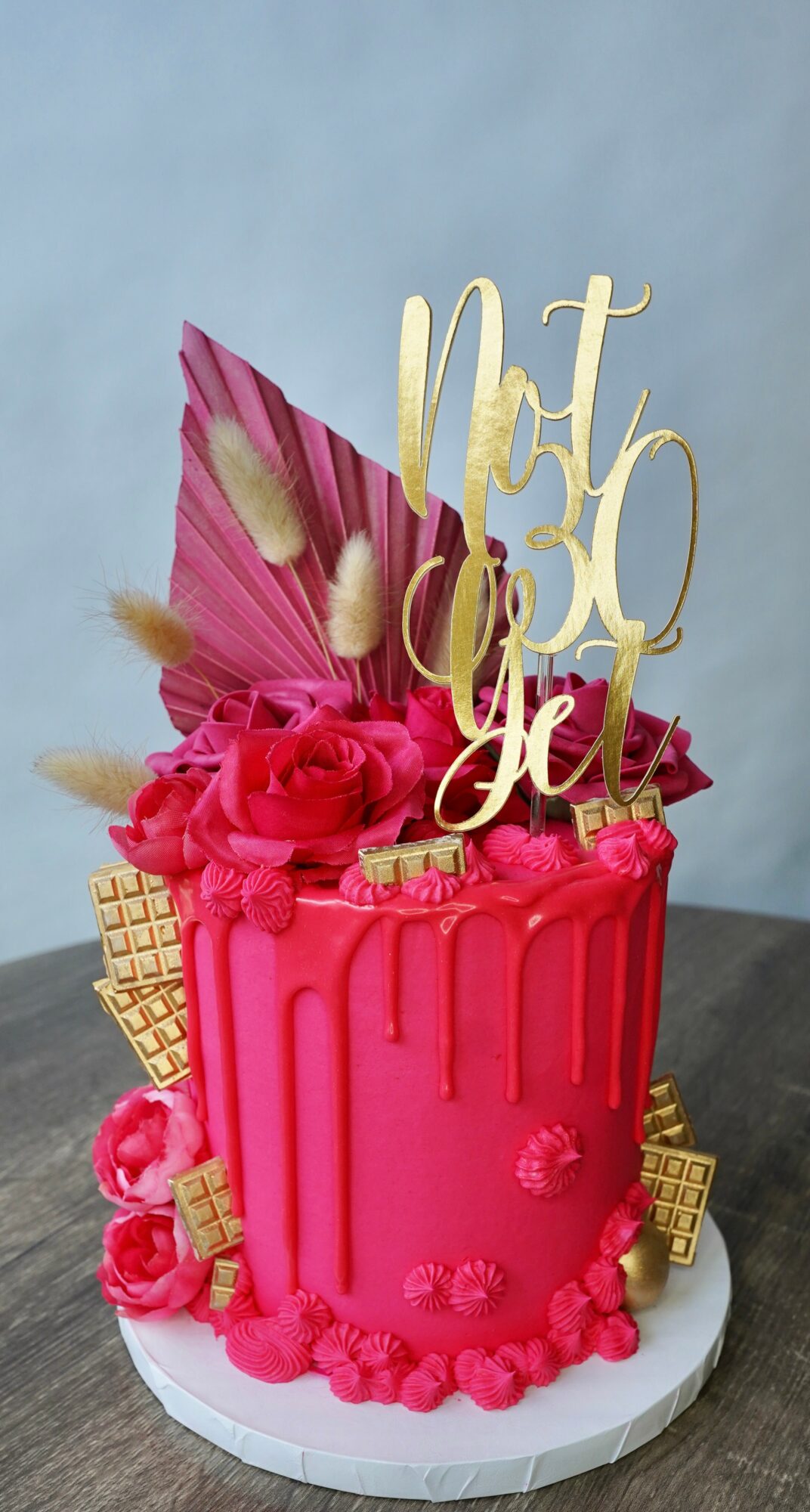 Pink birthday cake decorated with flowers, leaves, and a gold topper that says