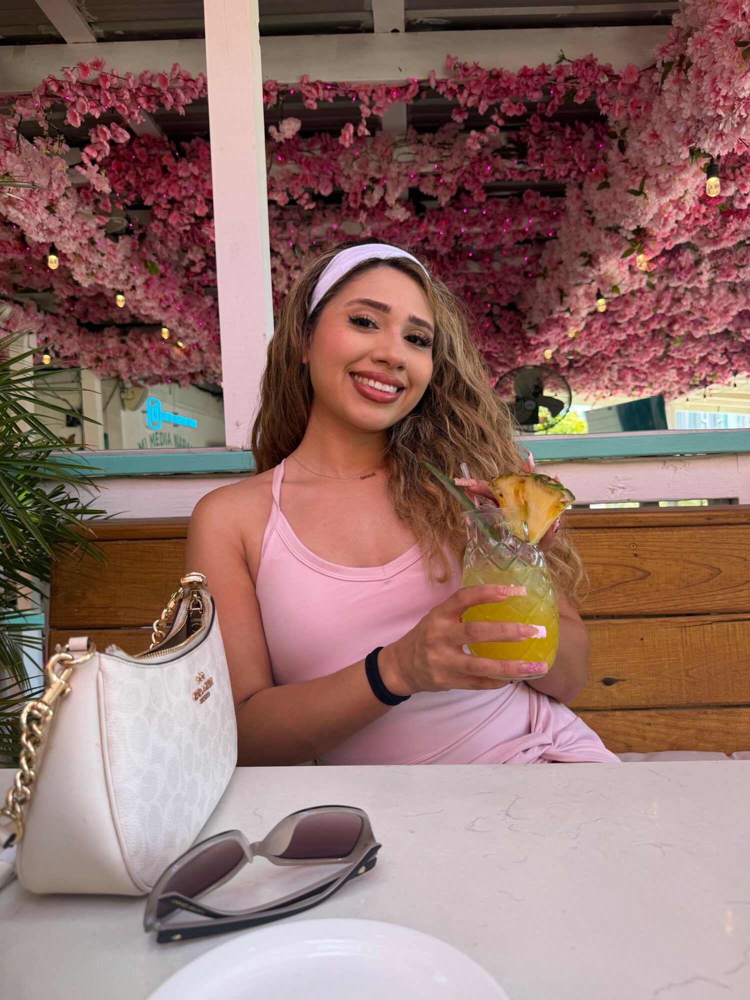 Young woman with long hair, wearing a pink top, smiling, holding a yellow drink, sitting at a table outdoors with pink flowers in background.