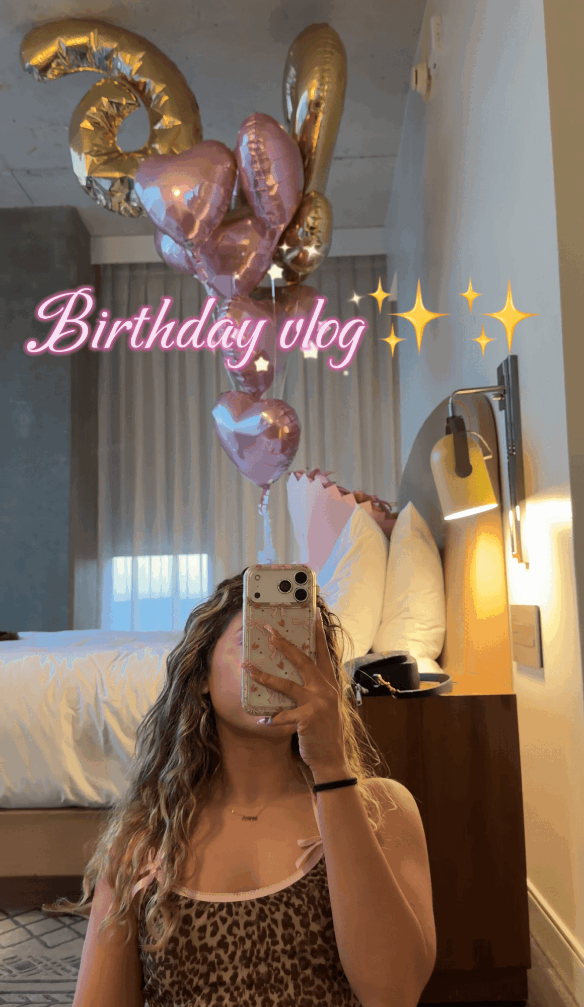 Person taking a mirror selfie in a hotel room with birthday balloons and a bed in the background.