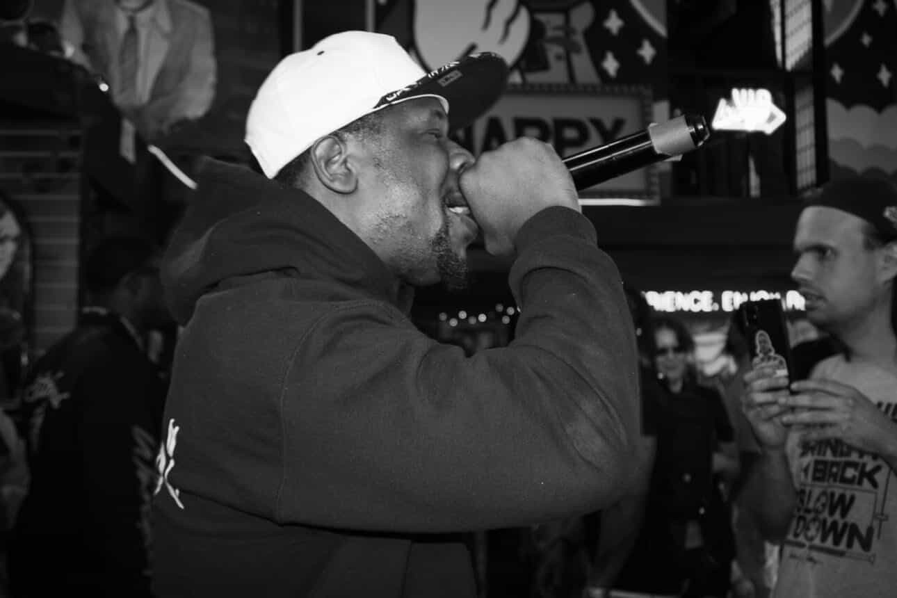 Person wearing a cap and hoodie singing into a microphone in a lively indoor setting with people around.