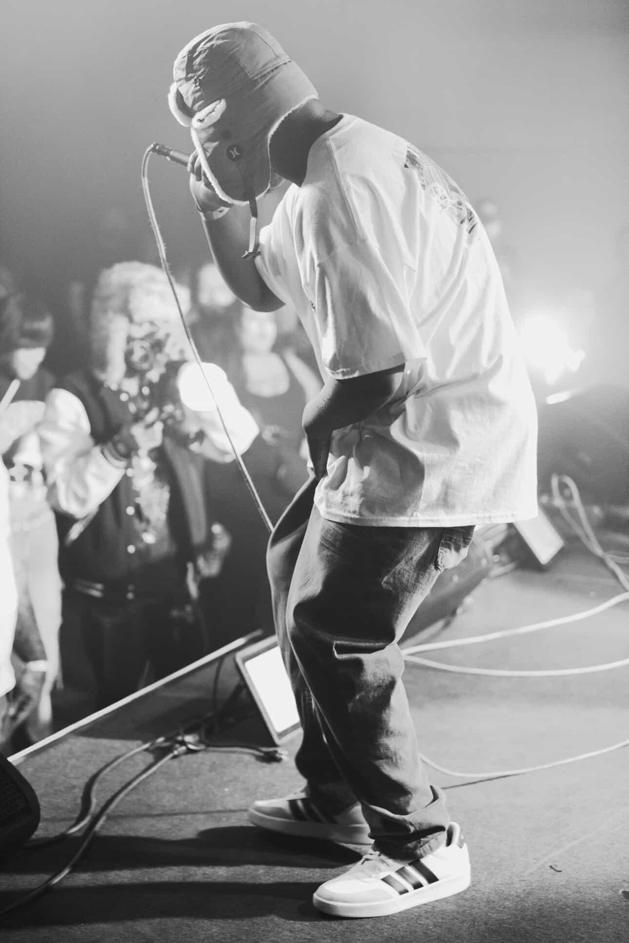 Performer on stage with microphone, audience in background, black and white photo, casual clothing, sneakers, performing passionately.