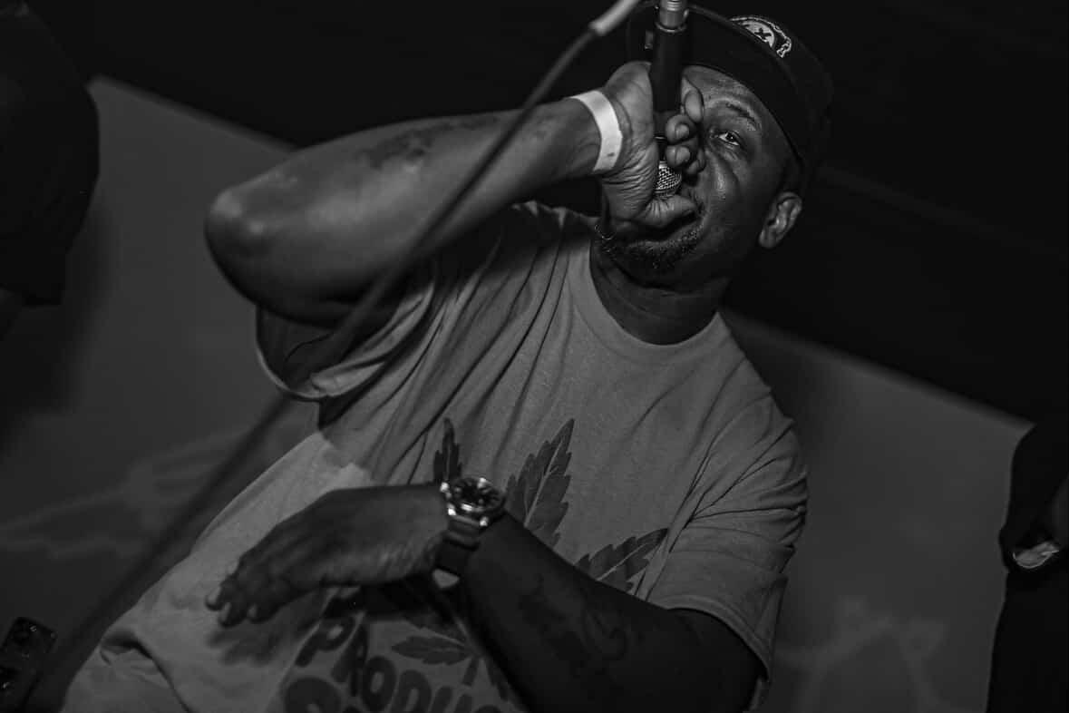 Person singing into a microphone with eyes closed, wearing a cap and casual clothing, in a dark setting.