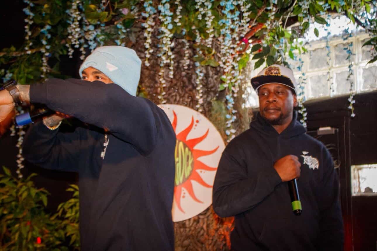 Two men stand outdoors under a flowering tree, one with a microphone, the other with a beanie, both in dark clothing.