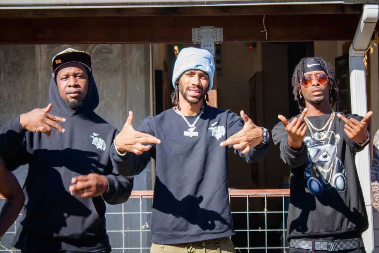 Three men standing outdoors, making hand gestures, wearing dark clothing and accessories, smiling and posing for the camera.