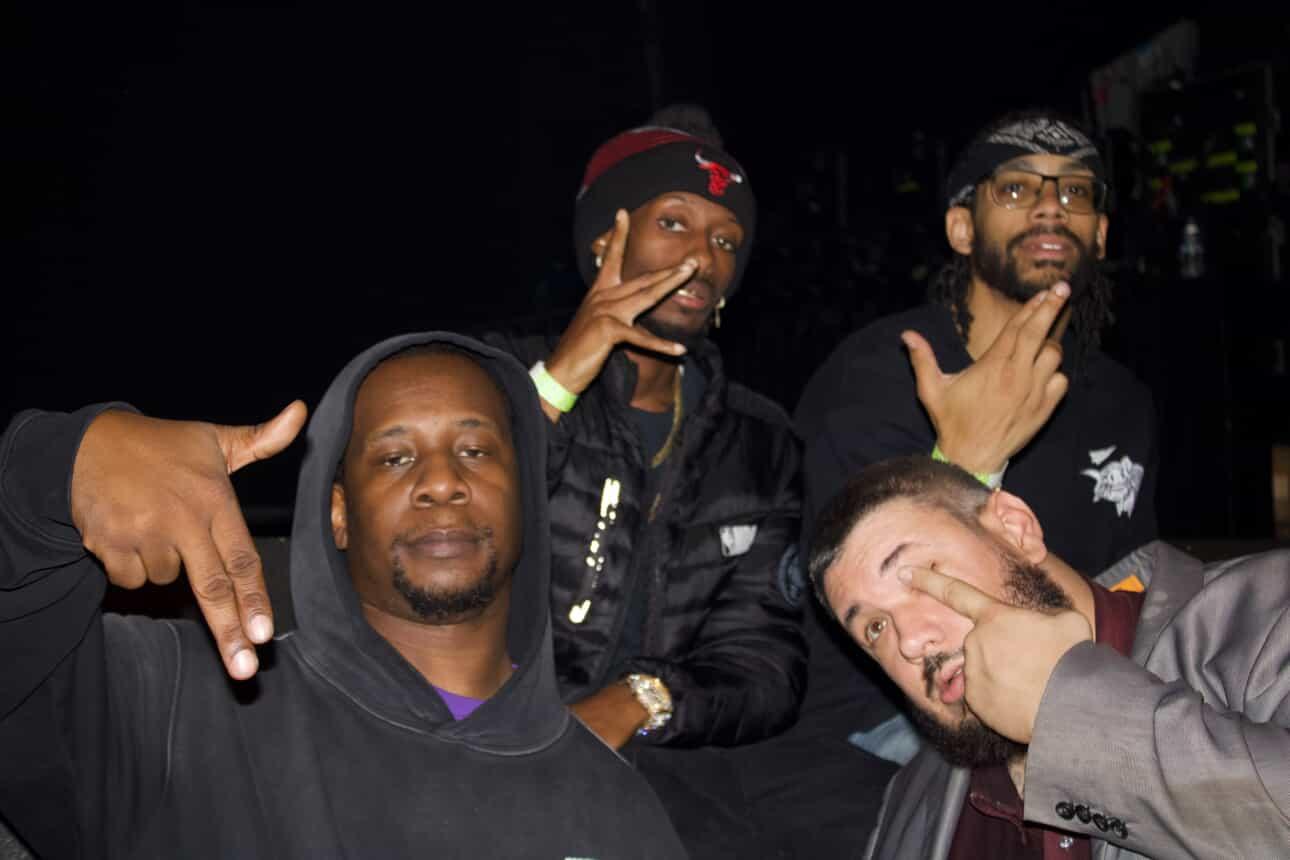 Four men posing together in a dark setting, some making hand gestures, one smiling widely, others with serious expressions.