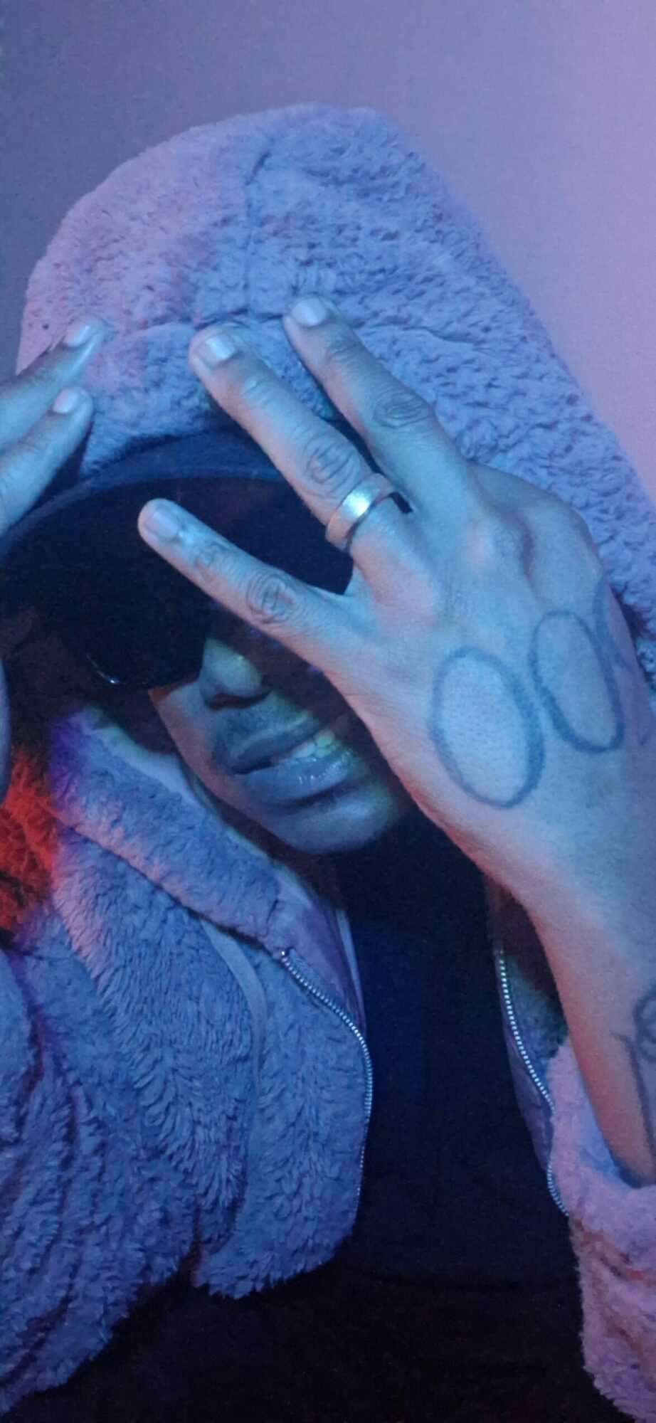 Person's hand with rings, tattoos, and rings on fingers, resting on a textured fabric surface.