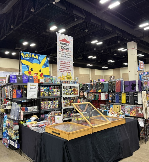 Booth with Pokémon Pikachu poster, shelves with toys and collectibles, and a person behind the table at an indoor event.